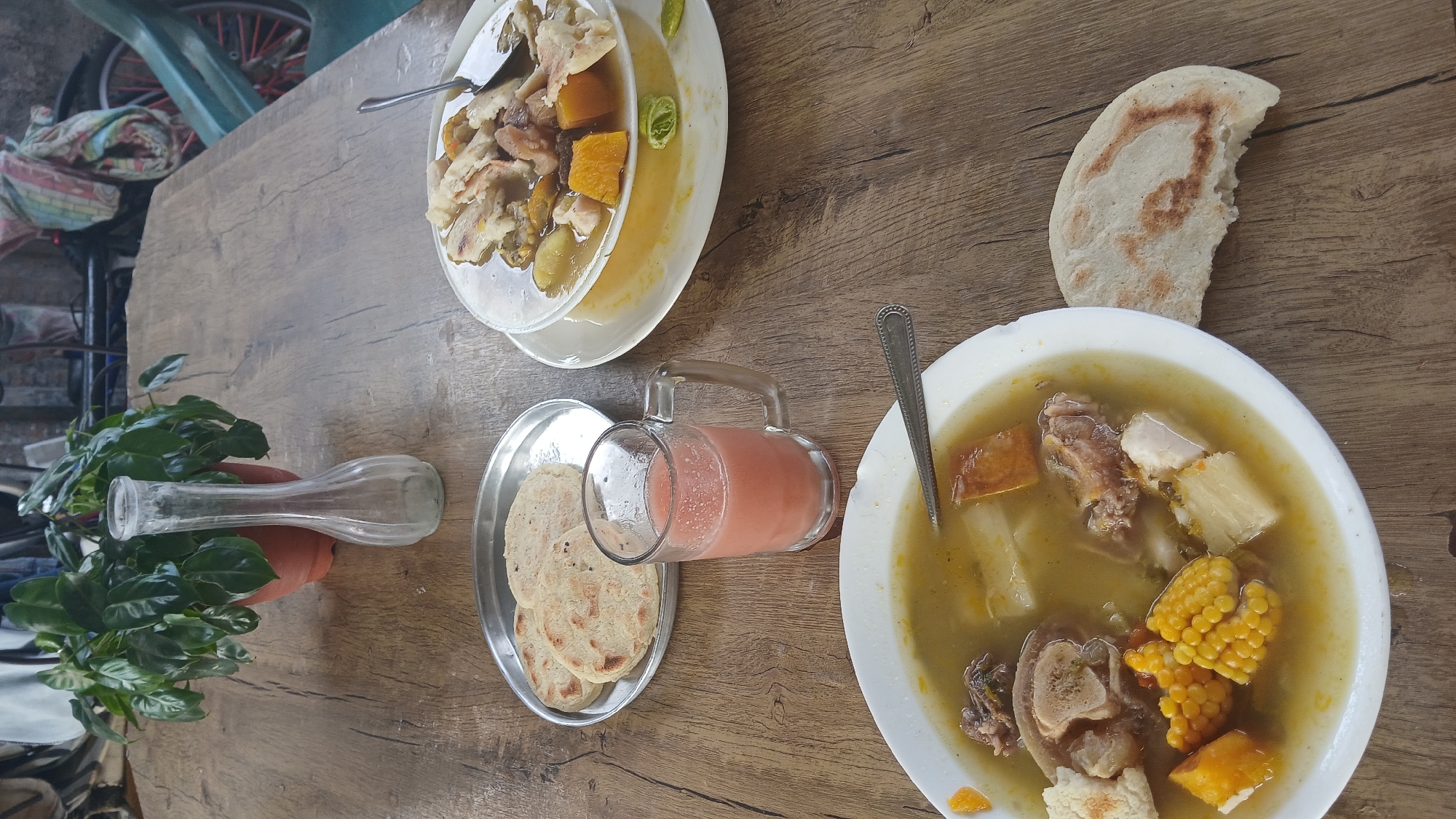 The image shows a wooden table in what appears to be a restaurant or food establishment. On the table, there are two bowls of soup with visible ingredients such as meat, corn, and squash. Beside one of the bowls is a piece of flatbread. A metal tray contains more flatbread and a glass pitcher filled with a pink beverage. To the side, there is a vase with greenery. In the background, parts of a bicycle can be seen. The location is identified as Altagracia, Venezuela.