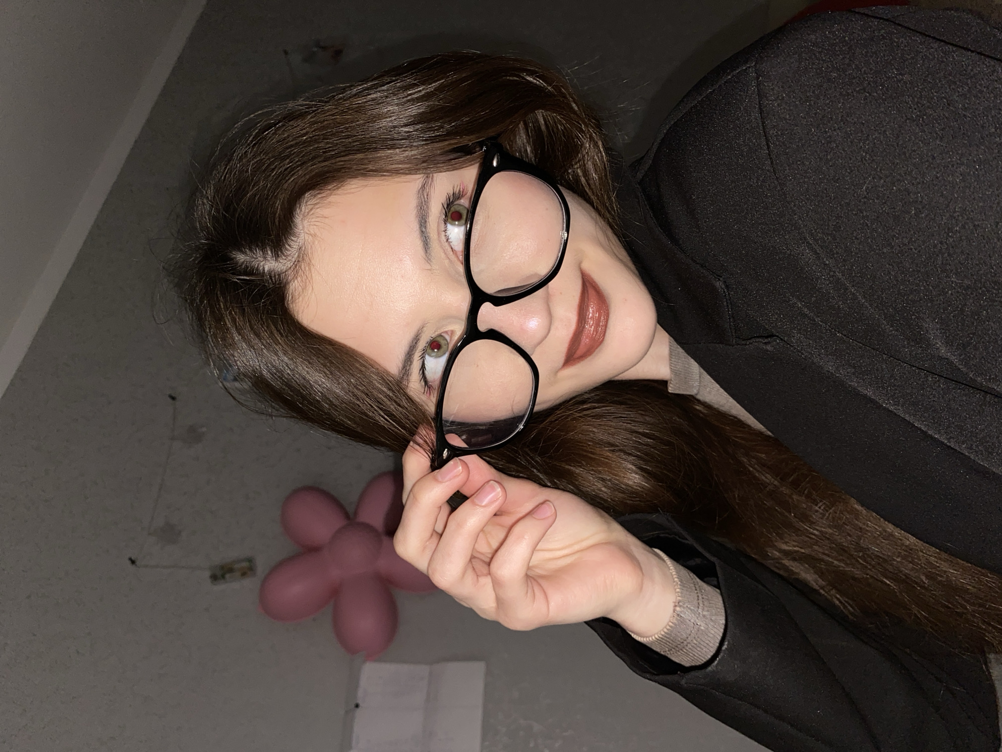 An angled portrait captures a young woman indoors, likely in Votkinsk, Russia. She has long, dark brown hair parted on the side, flowing past her shoulders. She wears prominent black-framed glasses, which she holds with her right hand. Her eyes, appearing brown or hazel, show distinct red reflections from the lighting as she looks upwards and slightly to the right. A subtle, gentle smile graces her lips, which are colored with brownish-pink lipstick.
She is dressed in a dark, possibly black, coat or jacket, with a lighter-colored top, possibly beige or light brown, visible at the cuff of her left arm. The background consists of a plain, light grey wall. To the lower left, a pink balloon shaped like a flower is visible. Faint marks are present on the wall, along with a small, rectangular object taped to it, though its exact nature is unclear. The lighting suggests an indoor setting, potentially in the evening or under artificial light, creating shadows and highlights. The overall expression on her face conveys a pleasant and slightly playful mood. No visible text is present in the image.