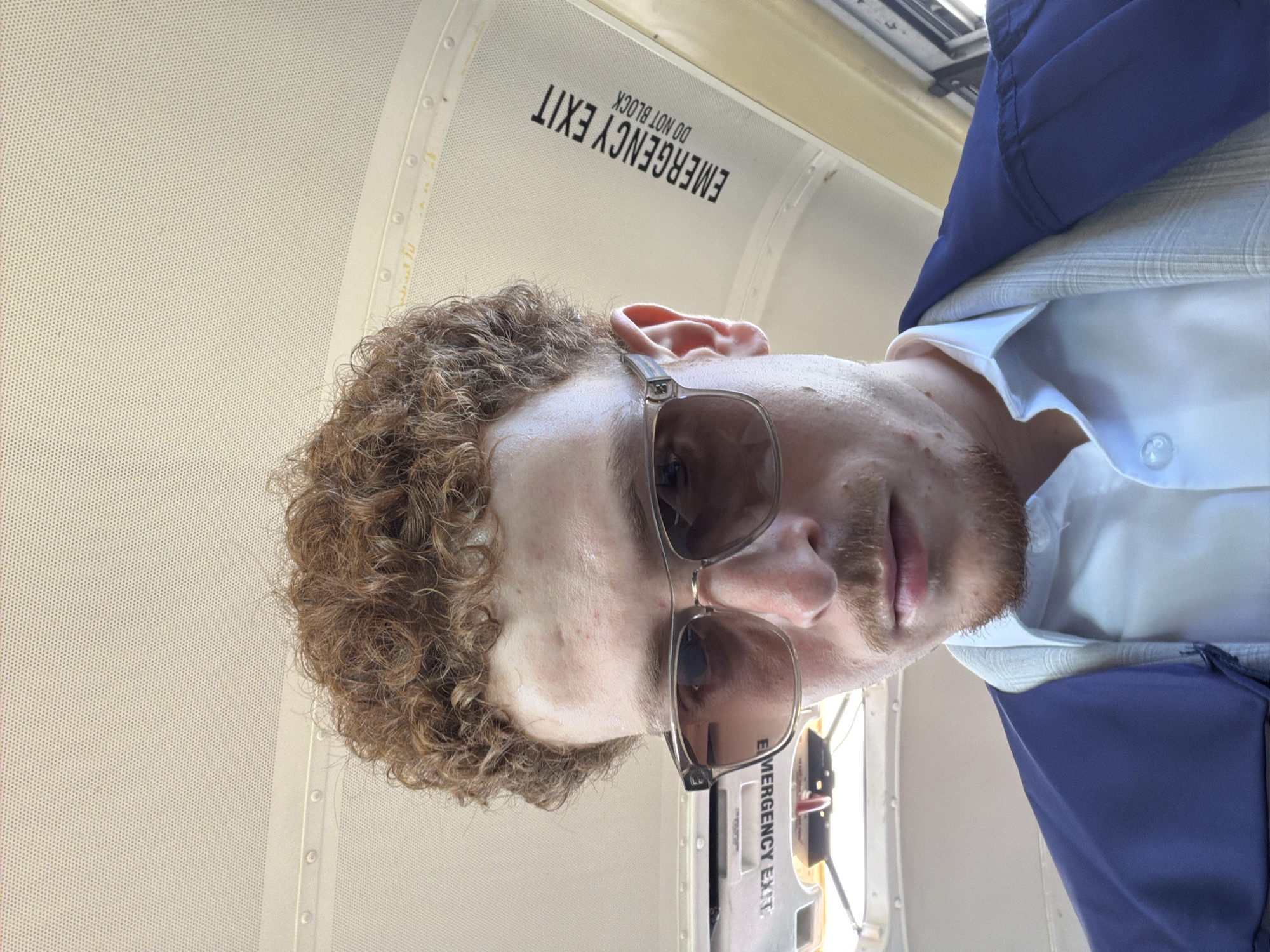 A Caucasian male wearing sunglasses, a light blue dress shirt with a button visible, a patterned light gray blazer, and a blue blazer is situated in what appears to be the interior of a bus or similar vehicle. The "EMERGENCY EXIT DO NOT BLOCK" sign can be seen above the individual. The specific location within the city of New York cannot be definitively determined from this image.