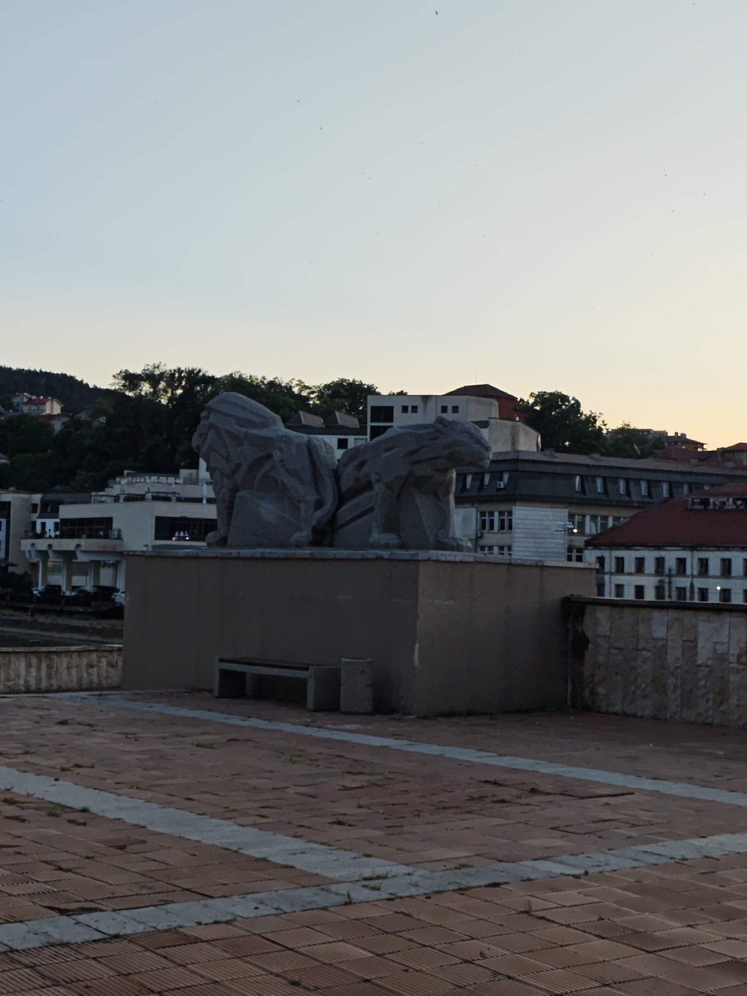 The image shows a monument of two stone lions, a bench, and a trash can in the foreground. Behind the monument, there are several buildings and trees. The sky is clear and the sun is setting. The monument is located in the city of Lovech, Bulgaria.