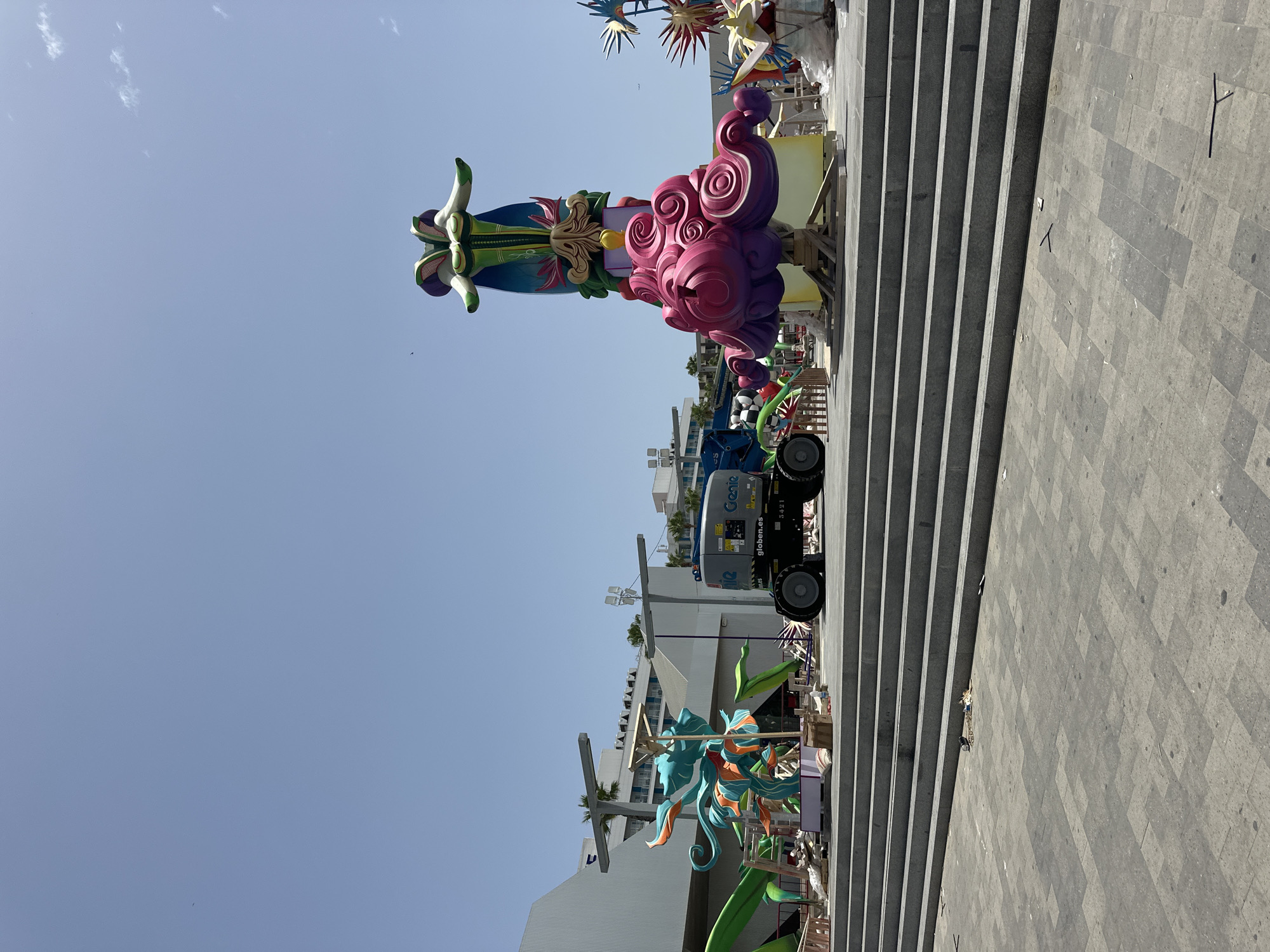 In an outdoor urban plaza, large, colorful, abstract sculptures are set up on a tiled surface. The sculptures are varied in design, with one resembling a stylized dragon in teal and orange, and another a tall, slender figure with a pink, cloud-like base and a green and purple top. A Genie brand aerial lift is positioned near the sculptures. The scene is set against a backdrop of a modern building with a white facade and a clear blue sky. The foreground features wide, grey stone steps leading up to the plaza. The overall impression is one of public art installation or preparation for an event.