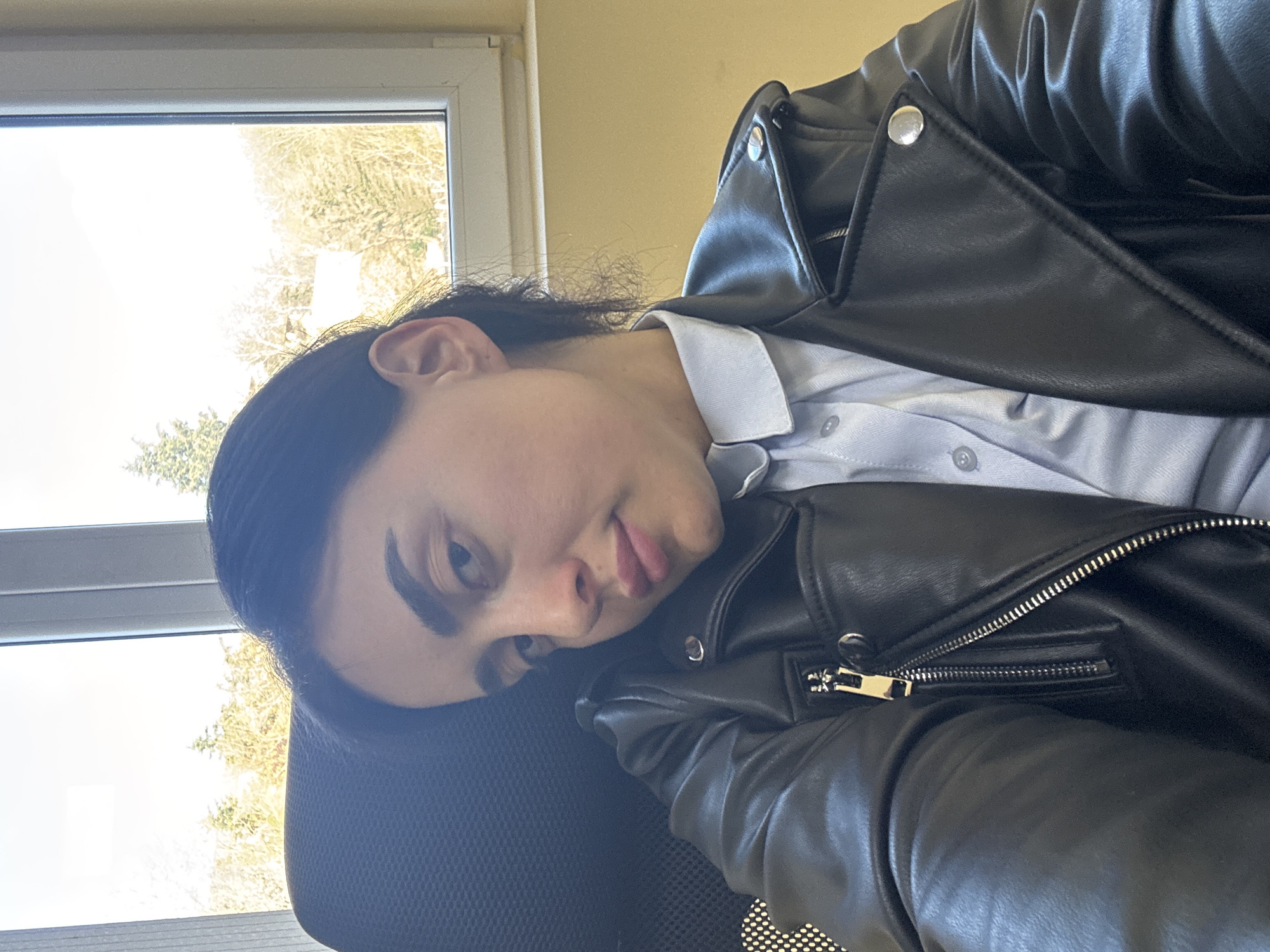 A person with dark hair is sitting in an office chair, wearing a light blue collared shirt and a black leather jacket. The person's face is turned to the left, with a pensive expression. A window is visible in the background, showing trees and sky. The location is presumed to be in Tbilisi, Georgia, but there are no visible clues in the image to confirm this.