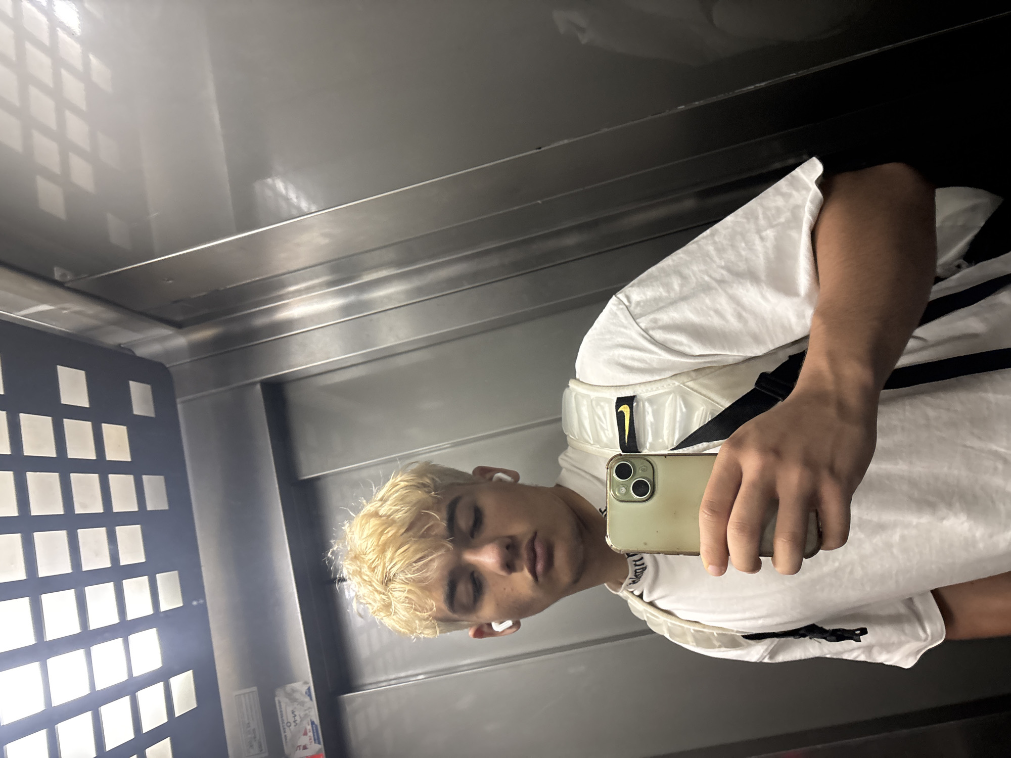 A young person with dyed blonde hair and white earbuds is taking a selfie in an elevator. They are wearing a white t-shirt and a reflective vest with a Nike logo. The elevator has a metal interior and a tiled ceiling. The image is taken in the city of Sofia, Bulgaria.