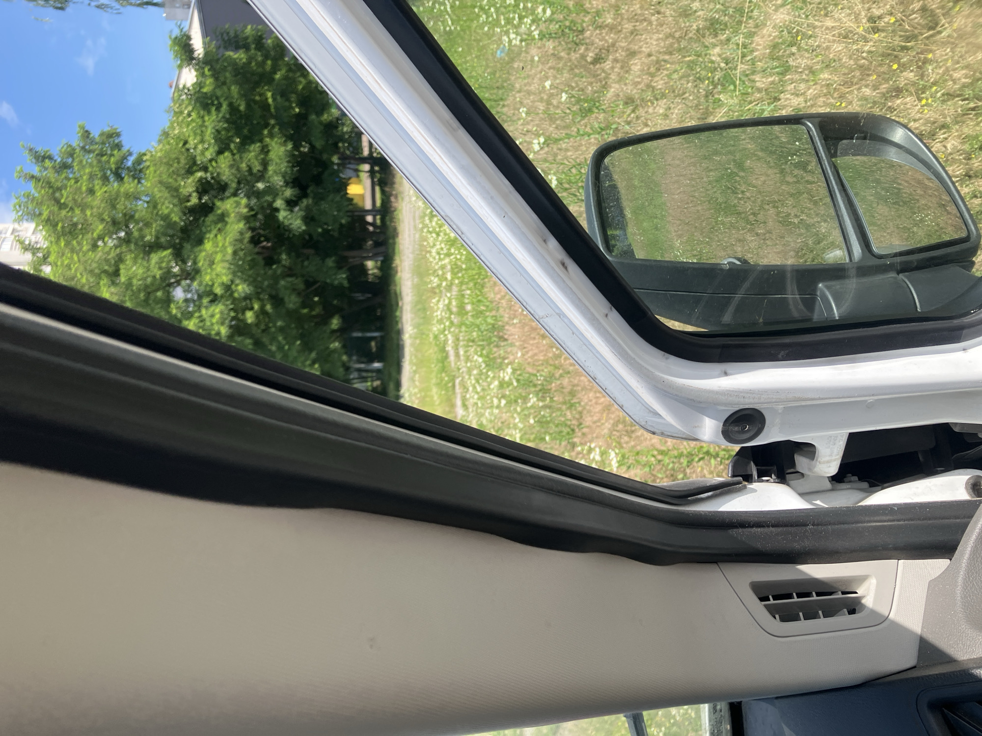 This media file depicts an exterior view of a white vehicle, likely a van, looking out from the driver's side door frame. The door is partially open, revealing the interior trim and a side mirror. Beyond the vehicle, a grassy area dotted with white wildflowers stretches into the background, suggesting an outdoor setting in a rural or suburban environment. Trees with lush green foliage are visible to the left, and a building can be seen behind them. The sky is blue with some clouds, indicating daytime and fair weather.
The image focuses on the details of the vehicle's door frame, including the weather stripping and the edge of the door. The side mirror shows a reflection of the surrounding grassy field and some faint images of the sky and trees. A small grey vent is visible on the interior paneling of the vehicle in the lower right corner of the frame.
The scene appears to be captured during daylight hours, given the bright sunlight and clear sky. The presence of wildflowers suggests a spring or summer season. The location context provided indicates Kyiv, Ukraine, which further contextualizes the greenery and common types of buildings. There is no discernible activity or event taking place; the image is a static snapshot of the vehicle's exterior and its immediate surroundings. No text is visible in the media file.