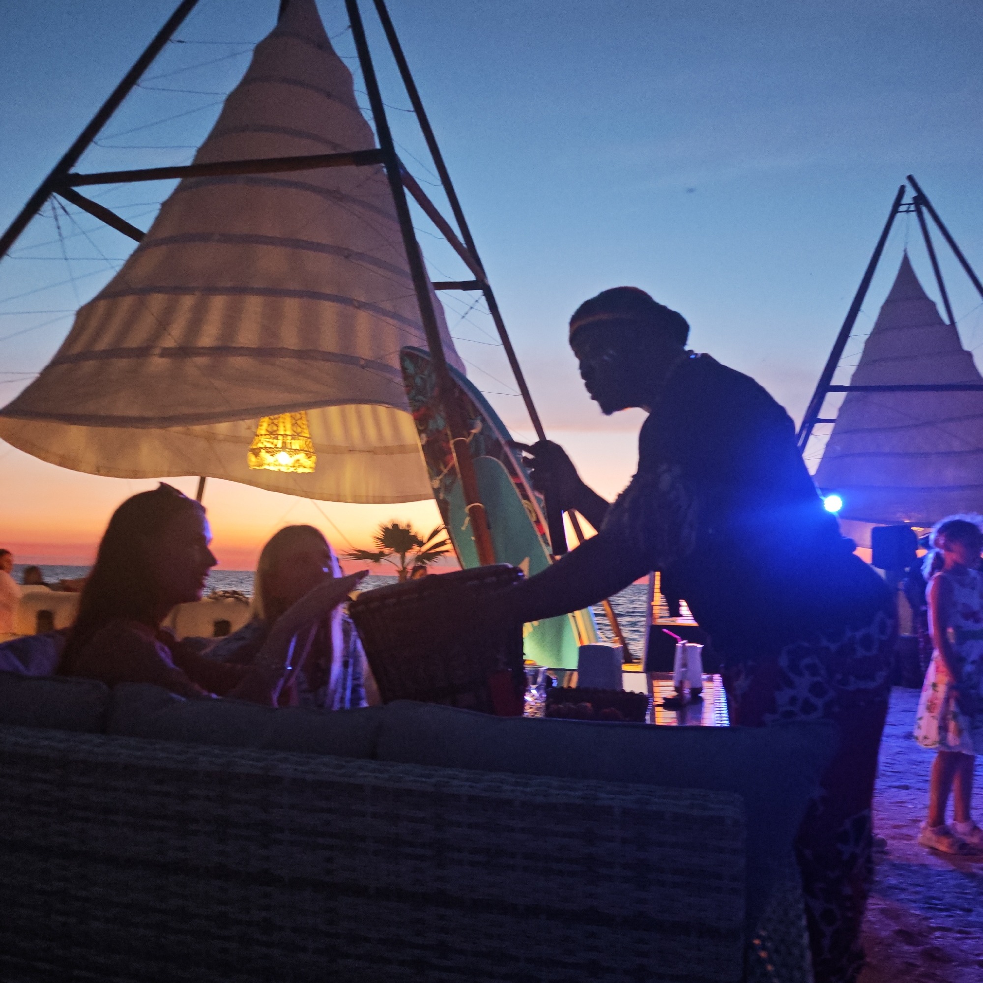 At dusk, on a beach in Мысовское сельское поселение, Ukraine, a group of people are gathered around a low table under a canopy with a conical tent-like structure. In the foreground, two women are seated on a sofa, silhouetted against the twilight sky. To their right, a person with their face painted and wearing a traditional outfit is standing and appears to be interacting with the group. Further to the right, a young girl in a floral dress is standing. The sea is visible in the background, with a warm sunset glow on the horizon. The scene is lit by ambient light from the setting sun and a decorative lamp hanging from the canopy.