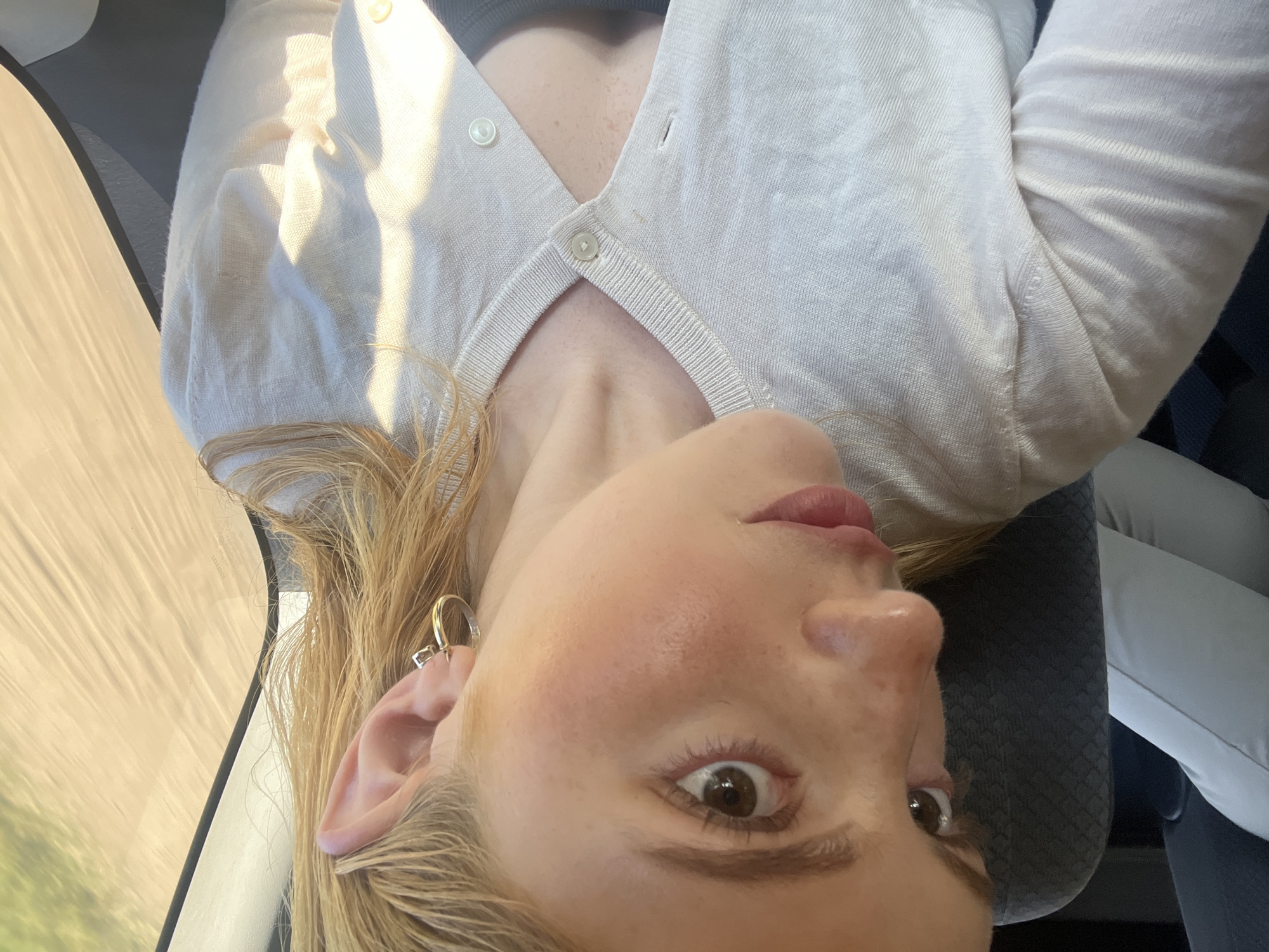 A close-up, rotated image depicts a fair-skinned woman with freckles and light blonde hair. She is wearing a cream-colored cardigan and has a gold earring. Part of a car window and a gray car seat are visible in the background. The location is not evident from the image.
