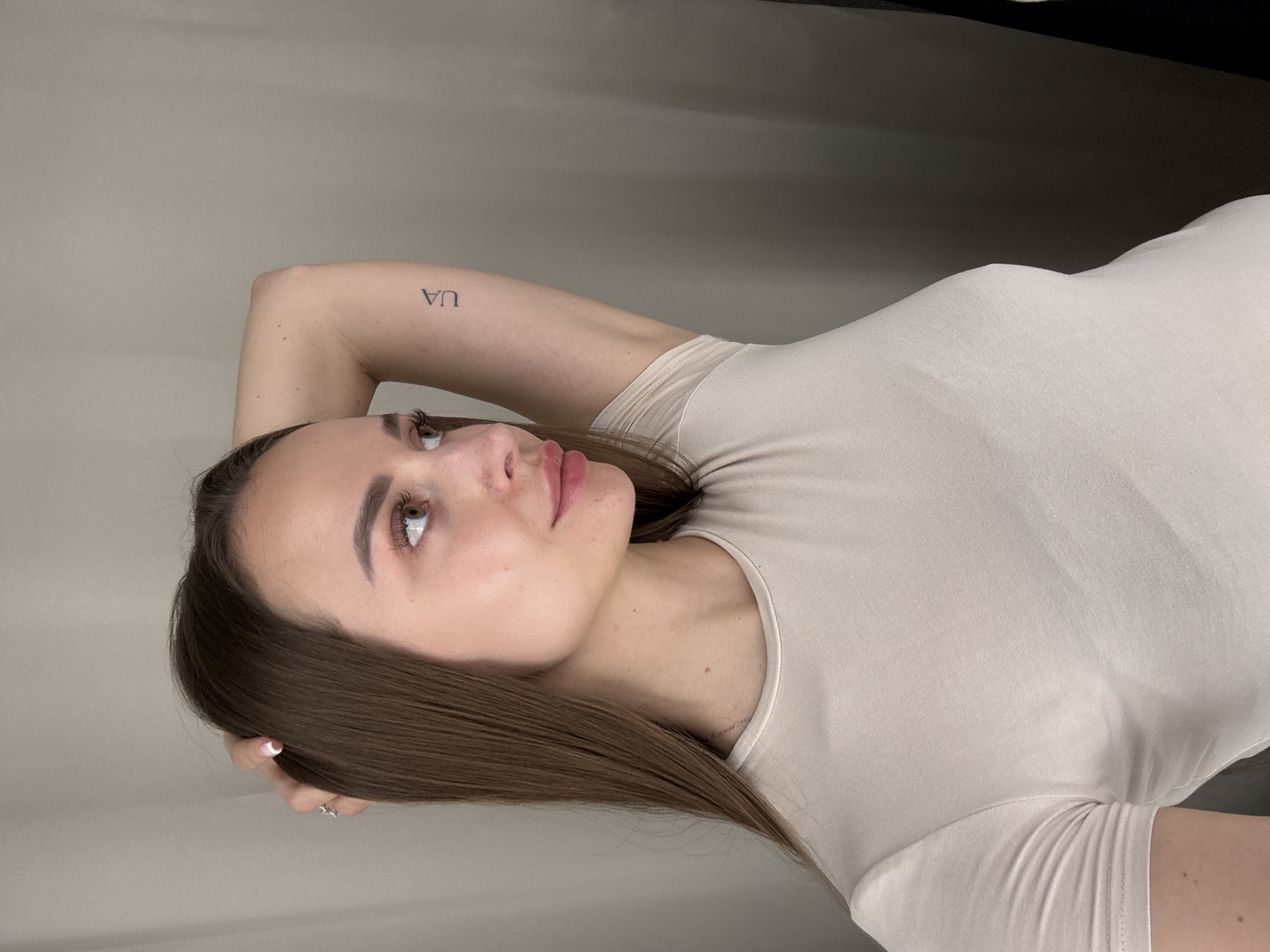 A woman with long, brown hair is reclining against a light-colored wall. She is wearing a light beige, short-sleeved shirt. Her left arm is bent behind her head. She has a small tattoo of the letters "UA" on her left arm. She is looking upwards. The location is unknown and cannot be determined from the image.