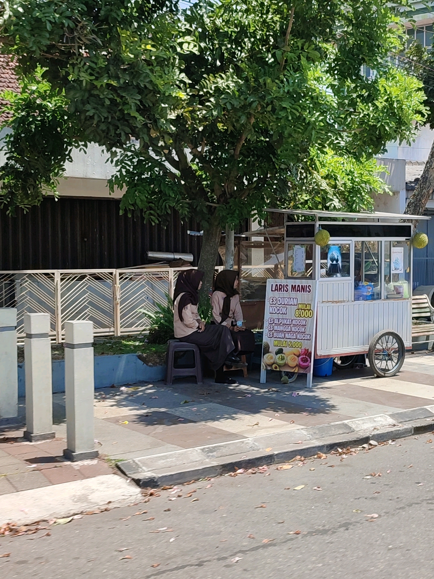 Two individuals wearing hijabs are seated on stools next to a mobile food cart. The cart displays the text "LARIS MANIS" and lists various ice cream flavors with prices. The food cart is white with a red trim and a wheel on the right side. Behind the cart, a tree with green foliage provides shade. In the background, there is a building with dark vertical panels and a decorative metal fence. The scene appears to be on a sidewalk next to a street, with fallen leaves scattered on the ground. The environment suggests a sunny day.