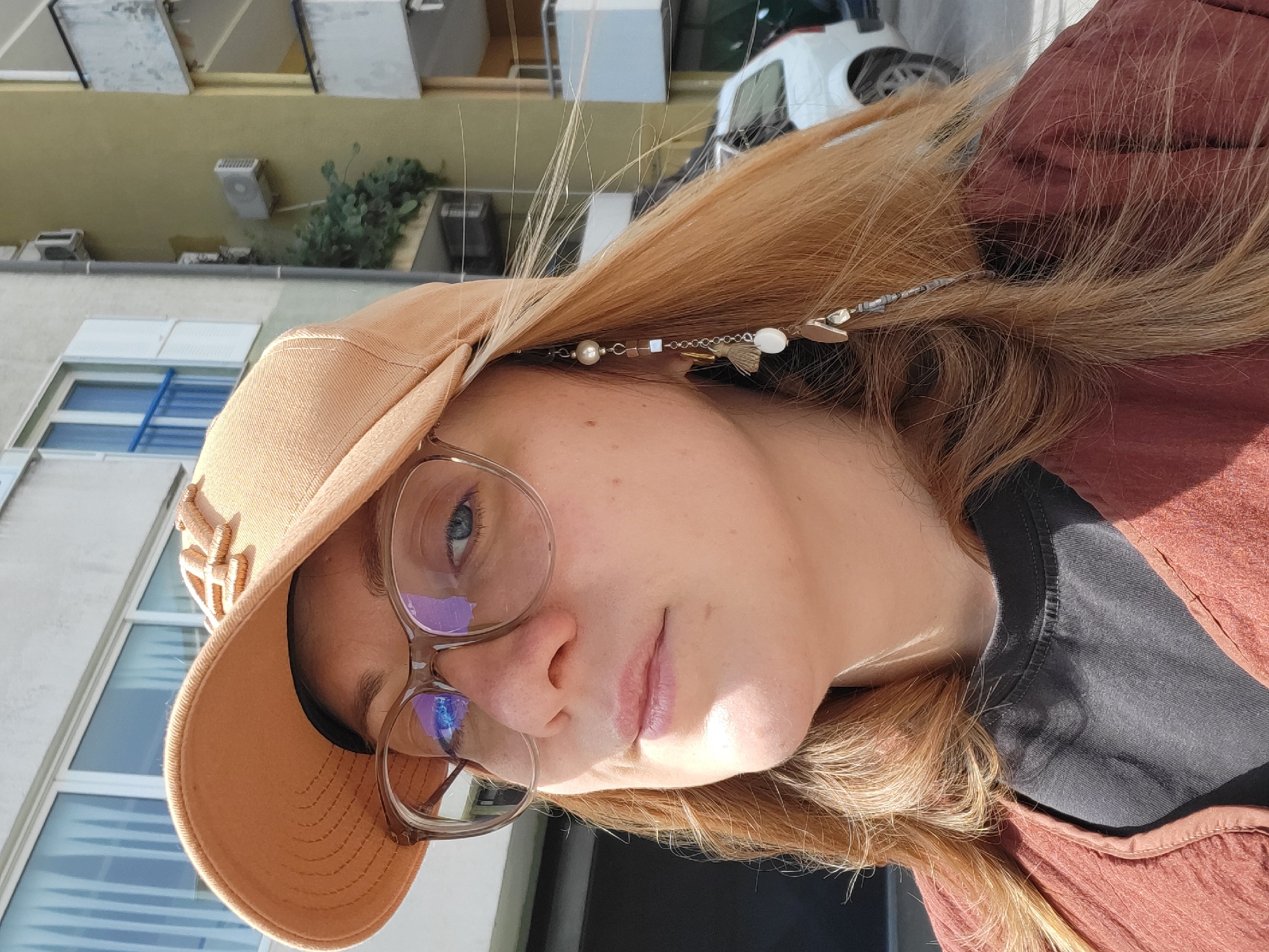 A medium close-up angled shot portrays a woman with blonde hair and blue eyes. She is wearing glasses and a tan-colored cap adorned with an embroidered wheat design. She also has a decorative beaded hair accessory. The woman's gaze is directed at the camera. She wears a dark shirt layered under a brown garment.
The background reveals elements of the built environment. The backdrop is a building with windows, some of which have blue frames, and a glimpse of greenery. A white car is partially visible in the upper right. Considering these details, the location appears to be urban. Although specific features are not present to confirm, it potentially could be Budva, Montenegro.