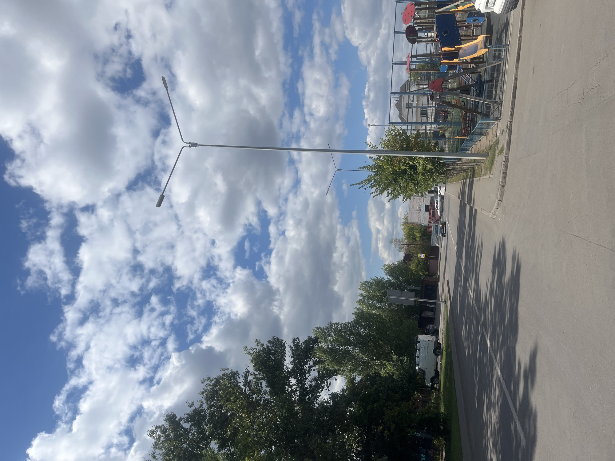 The image depicts a street scene on a partly cloudy day. On the left side of the street, there are lush green trees casting shadows onto the asphalt road. Several vehicles, including a white van and a black car, are parked or driving on the road. A sidewalk runs alongside the street, and beyond that is a fenced-in playground with various structures like slides and swings. In the background, there are buildings and more trees. The sky is a vibrant blue with scattered white clouds, suggesting fair weather. There are streetlights visible along the road. The overall scene appears to be a residential or suburban area.