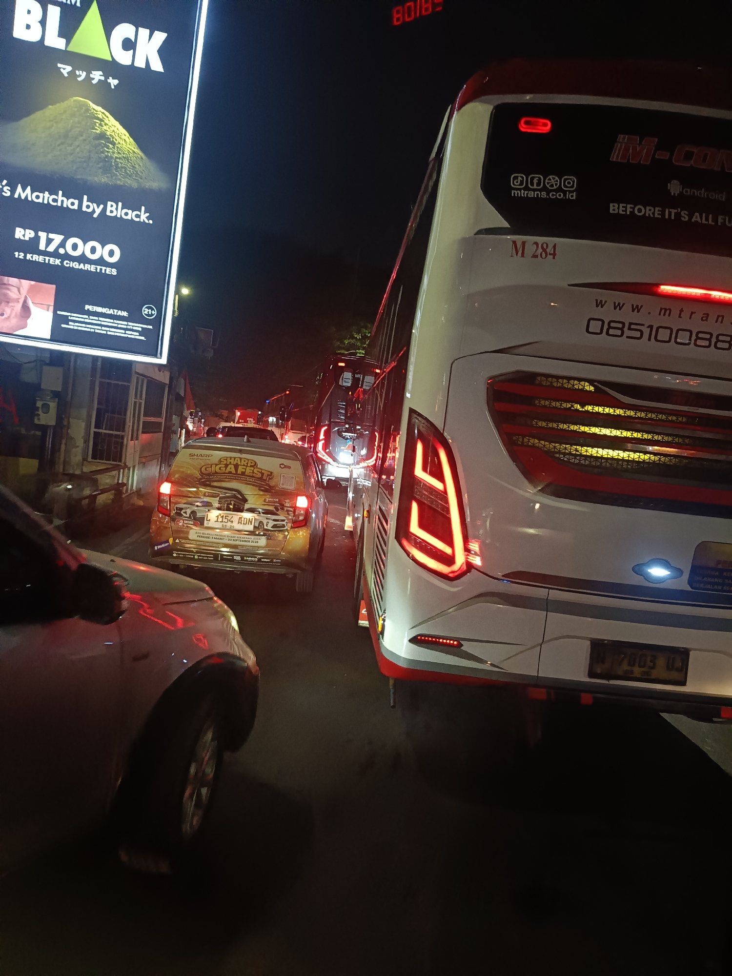 This image captures a nighttime scene of heavy traffic in Medaeng, Indonesia. The foreground is dominated by the rear of a white bus with red accents and distinctive taillights. Text on the bus indicates it is "IM-CON" and provides a website "www.mtran" and a phone number "08510088". Below this, a license plate reads "N 7003 UJ".
To the left of the bus, a smaller car is visible, adorned with a large graphic promoting "SHARP GIGA FEST" with a date of "30 SEPTEMBER 2020". The car's license plate reads "L 1154 ADN". Further left, a large illuminated billboard advertises "BLACK" cigarettes with "Matcha" flavor. Text on the billboard includes "RP 17.000" and "12 KRETEK CIGARETTES", along with a warning symbol "(21+) PERINGATAN".
The overall impression is one of a typical congested urban environment at night, likely in Indonesia given the visible license plate formats and language on the billboard. The bright lights of the vehicles and the billboard contrast with the dark night sky. No people are clearly discernible, but the presence of the vehicles and advertising suggests a public road or highway.