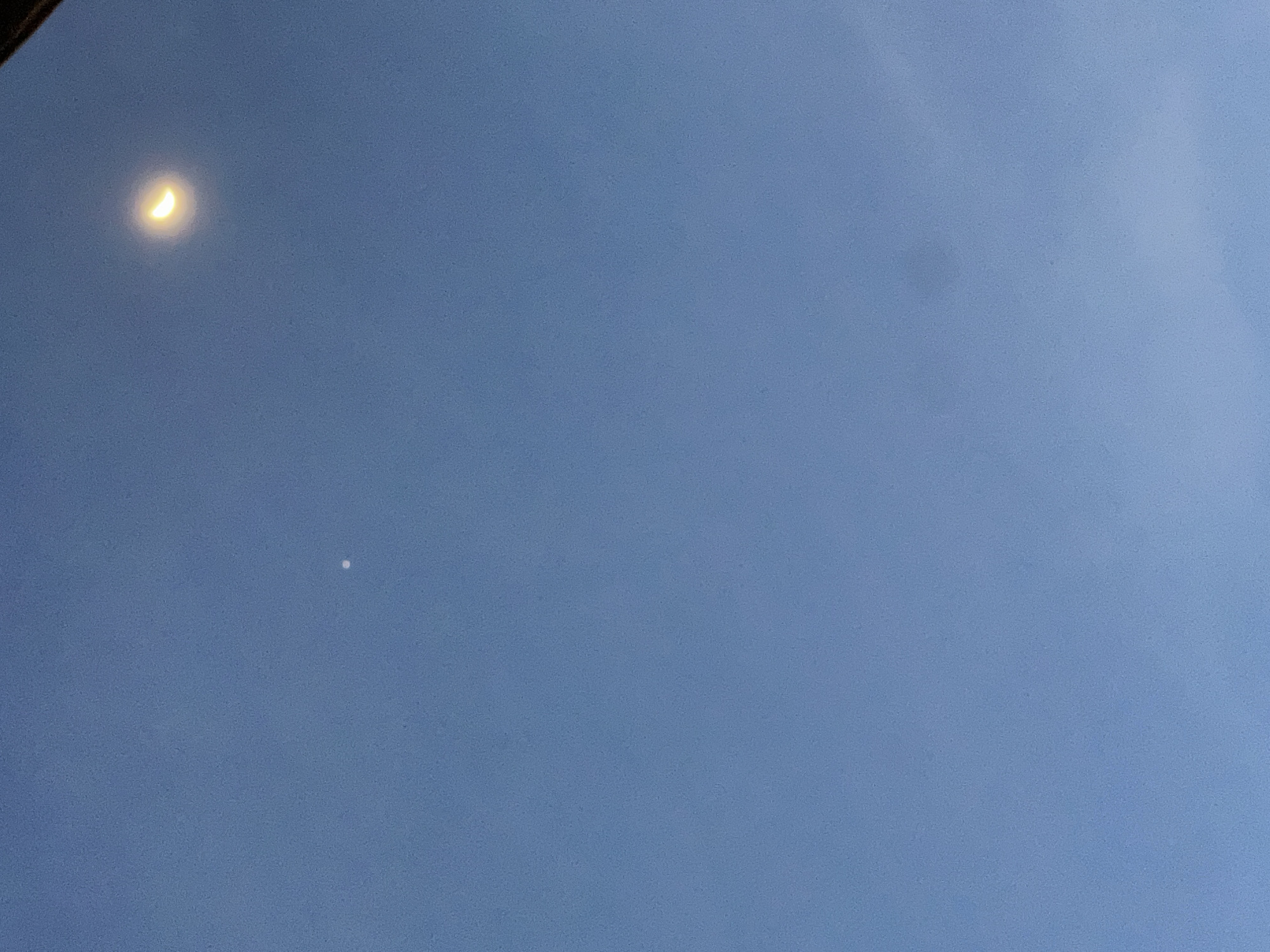 The image displays a clear blue sky, suggesting either daytime or twilight conditions. In the upper left portion of the frame, a distinct, bright crescent moon is visible, emitting a yellowish glow. Further down and to the right of the moon, a small, singular white dot is present, likely a planet such as Venus, appearing as another celestial body against the blue expanse. The sky is uniform in color, indicating clear weather without visible clouds. No people, other objects, or specific activities are depicted in the scene. There is no visible text in the image.