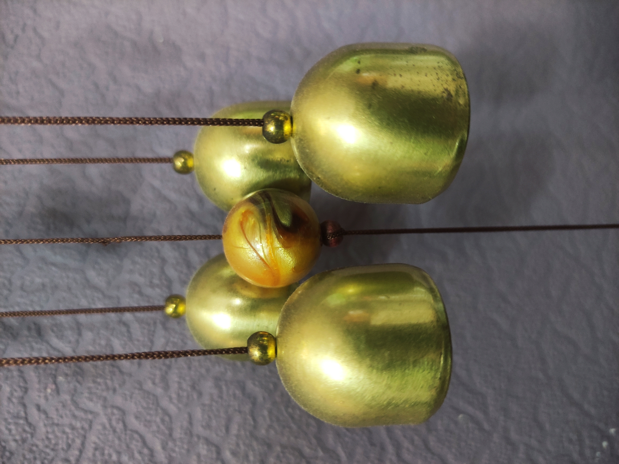 A close-up, slightly rotated, indoor shot depicts a wind chime. The chime consists of two sets of two tarnished gold-colored bells suspended by brown cords. Each bell is separated by small gold-colored beads. Centrally positioned between the two sets of bells is a marble or similarly patterned round object. The backdrop is a textured, gray surface. The location and city are unknown.