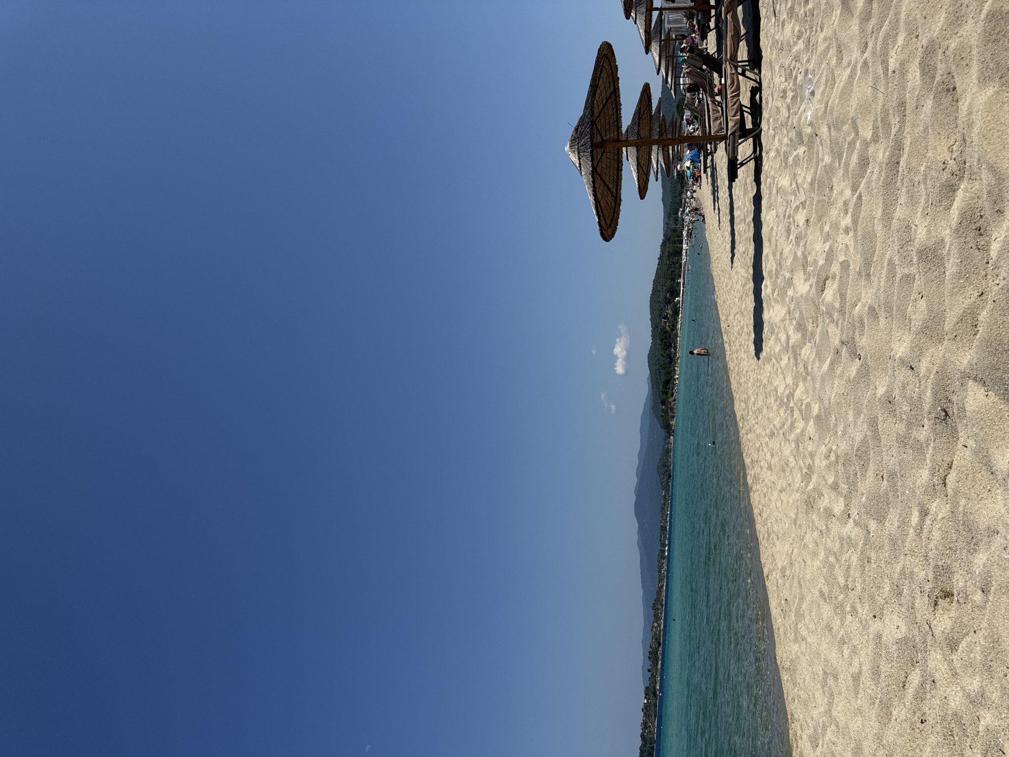 The image depicts a wide, sandy beach on a sunny day in Sithonia Municipal Unit, Greece. The clear, turquoise water of the Aegean Sea gently laps the shore. In the foreground, the sand is textured with ripples from the wind. Several thatched umbrellas stand in rows on the beach, casting shadows on the sand, with empty loungers beneath them. In the distance, a line of lush green hills forms the coastline. The sky is a vibrant, cloudless blue, with a sliver of the moon visible in the upper left quadrant. A few people are scattered along the shoreline, some swimming in the water and others relaxing on the beach. A lone individual stands waist-deep in the sea. The scene is peaceful and idyllic, conveying a sense of relaxation and summer vacation.