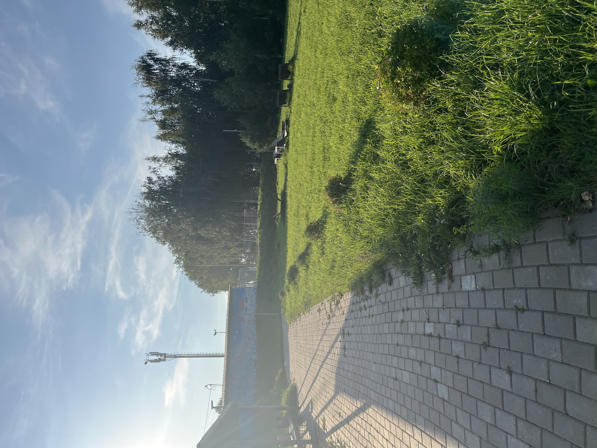 The image depicts a paved walkway in Ufa, Russia, leading towards a grassy area with scattered benches. A cell tower stands tall in the background, against a bright blue sky with wispy clouds. The walkway is lined with lush green grass and a few bushes. A mural adorns a building wall in the middle ground, featuring the text "С Днём Победы" (Happy Victory Day) and the dates "1941-1945". The sun casts long shadows across the scene, indicating it is either early morning or late afternoon. The overall atmosphere is peaceful and serene, suggesting a public park or recreational area.