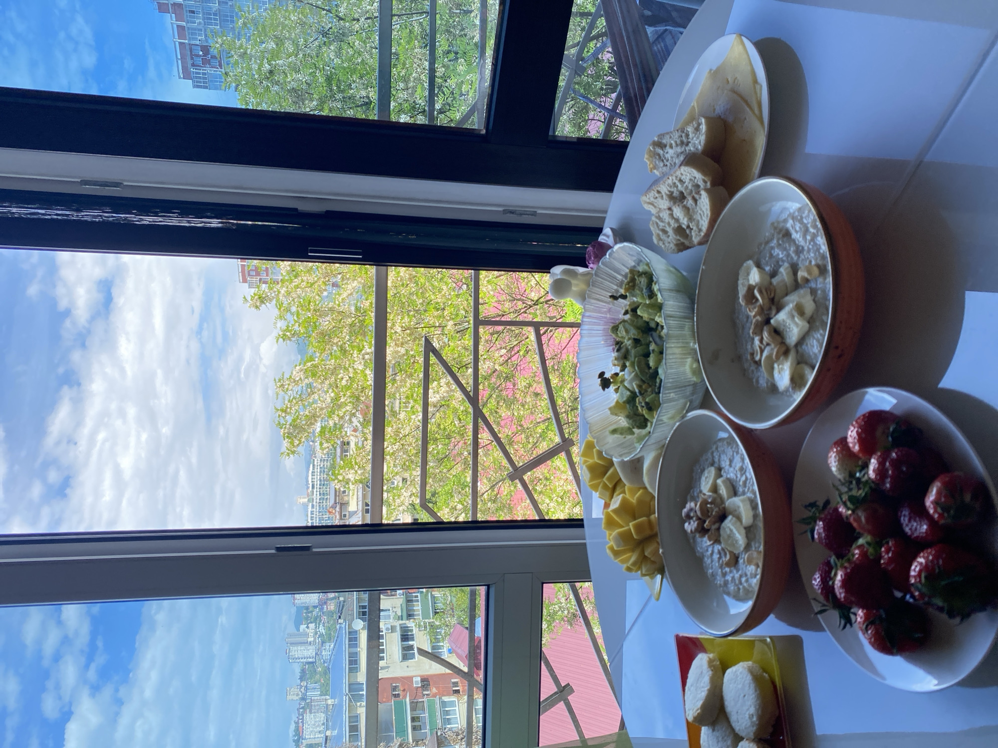 A table next to a window displays food items. There are two bowls of what appears to be oatmeal, topped with bananas and nuts. A plate holds a serving of strawberries, while another contains cookies. Additional plates include cheese and bread, as well as a green salad. Outside the window, urban buildings can be seen; this is Sochi, Russia. The day is bright, with blue skies and some clouds. Tree foliage is visible in the foreground of the window view.