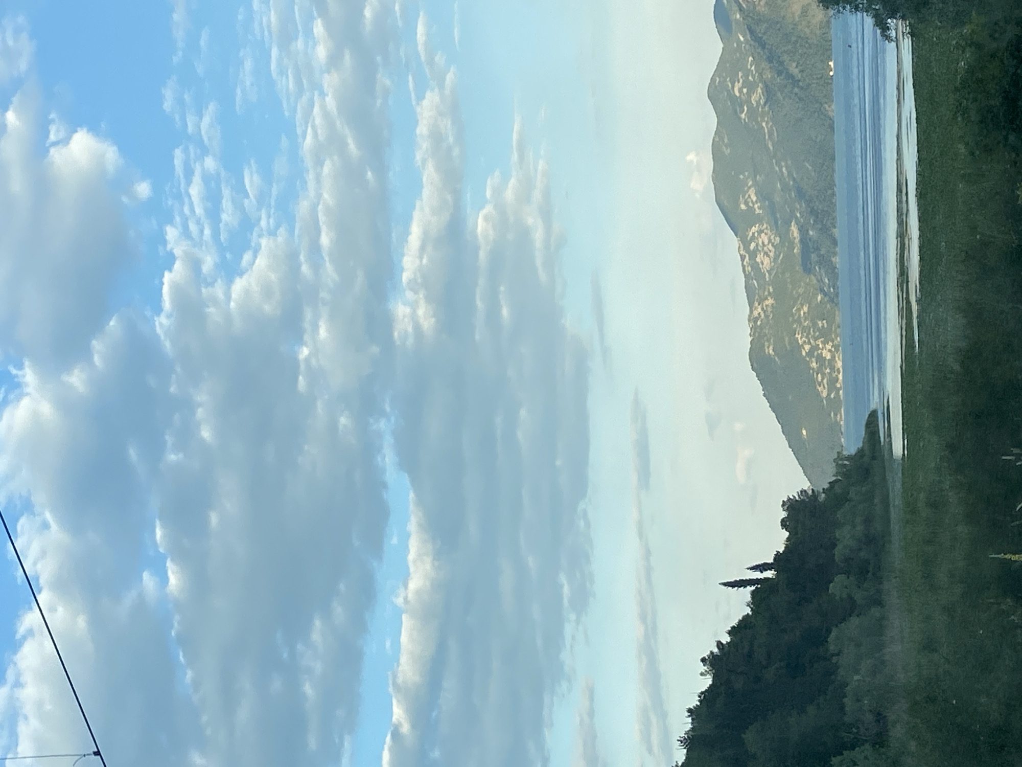 The image depicts a landscape with a cloudy sky dominating the left side. To the right, a body of water is bordered by mountainous terrain and a dense treeline. This scene is likely in Braćeni, Montenegro.