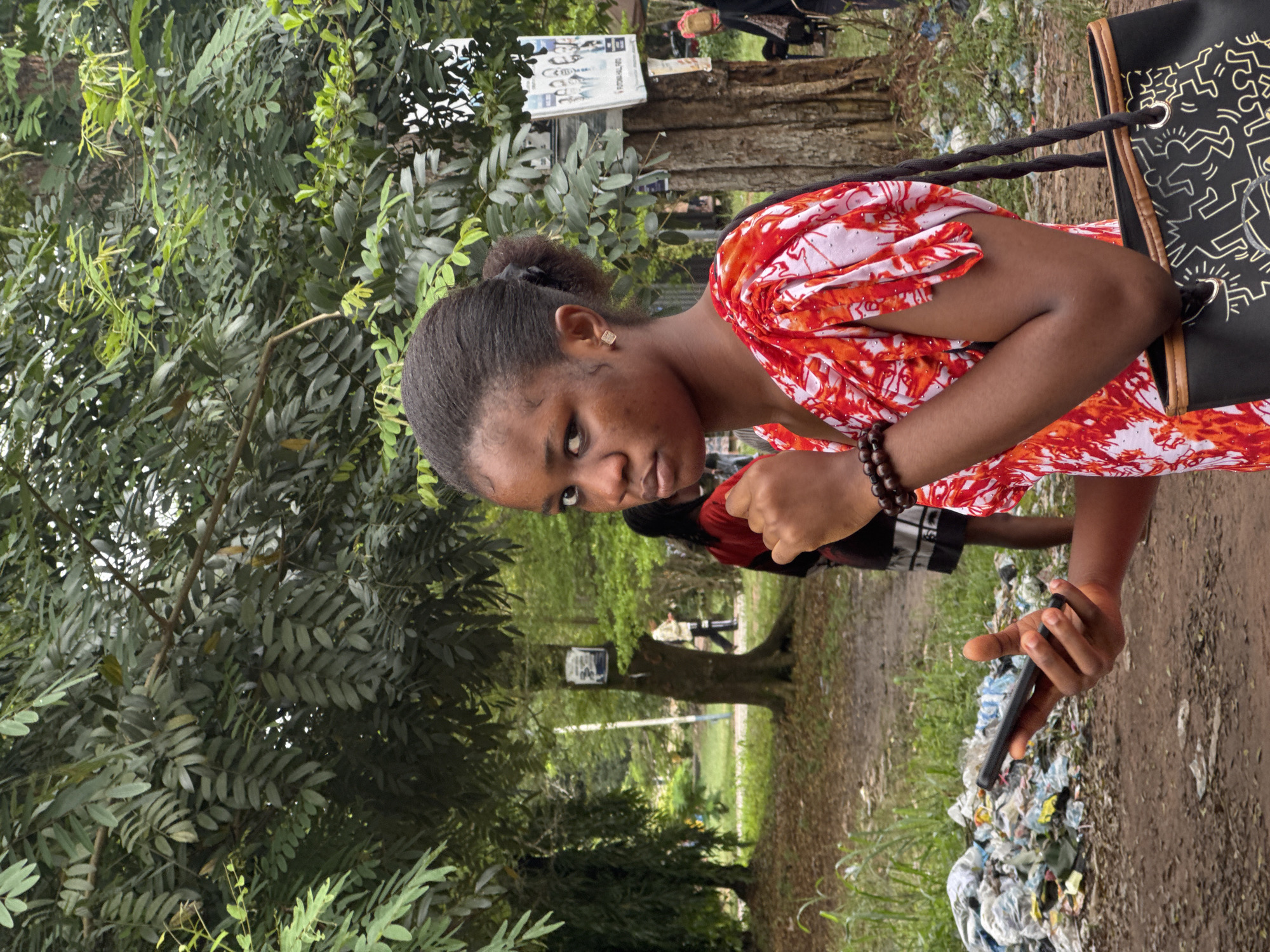 In Ihiagwa, Nigeria, a young Black female stands in what appears to be an outdoor, possibly unpaved area with abundant vegetation. She is facing the camera, looking directly at the viewer with a neutral expression. She has dark hair pulled back into a bun and is wearing a vibrant red and white patterned dress with flared sleeves. She is holding a black smartphone in her right hand, with her thumb on the screen. On her left wrist, she wears a dark beaded bracelet. A black bag with a Keith Haring-esque pattern is slung over her right shoulder.
The background shows trees and foliage, suggesting a park or natural setting within the city. There is a pile of plastic waste and debris to the left of the subject, indicating a potential environmental concern or an area of urban decay. In the middle ground, other people are visible, including a person in a red top and another in a light-colored jacket, walking away from the camera. A tree trunk with a poster attached to it stands behind the subject. The ground is a mix of dirt and scattered leaves. The overall lighting suggests an overcast or shaded day.