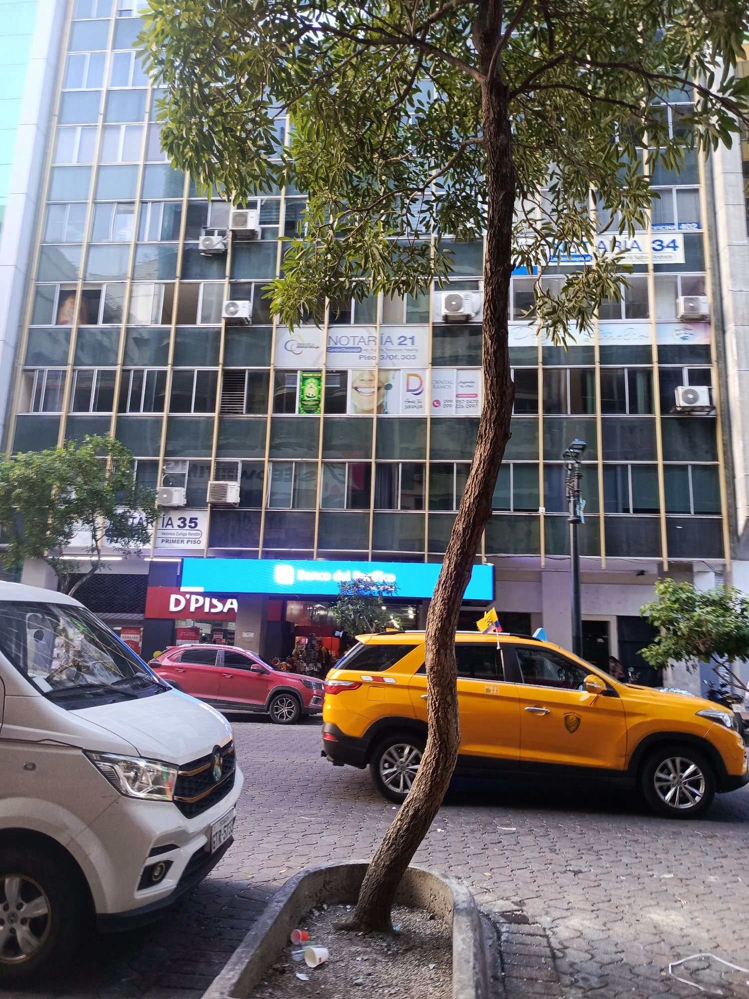 In Guayaquil, Ecuador, on a city street, a white van is parked on the left side of the frame, with its front facing towards the viewer and slightly to the right. To the right of the van, a yellow taxi with a national flag on its roof is parked. The taxi has its driver's side door visible, and it appears to be occupied. In the background, a multi-story building with many windows and air conditioning units is visible. Several signs are affixed to the building, including one for "NOTARIA 21" and "D'PISA". The street is paved with cobblestones, and there is a tree trunk in the foreground, partially obscuring the view of the building. Litter is present on the ground near the tree. The sky is overcast.
