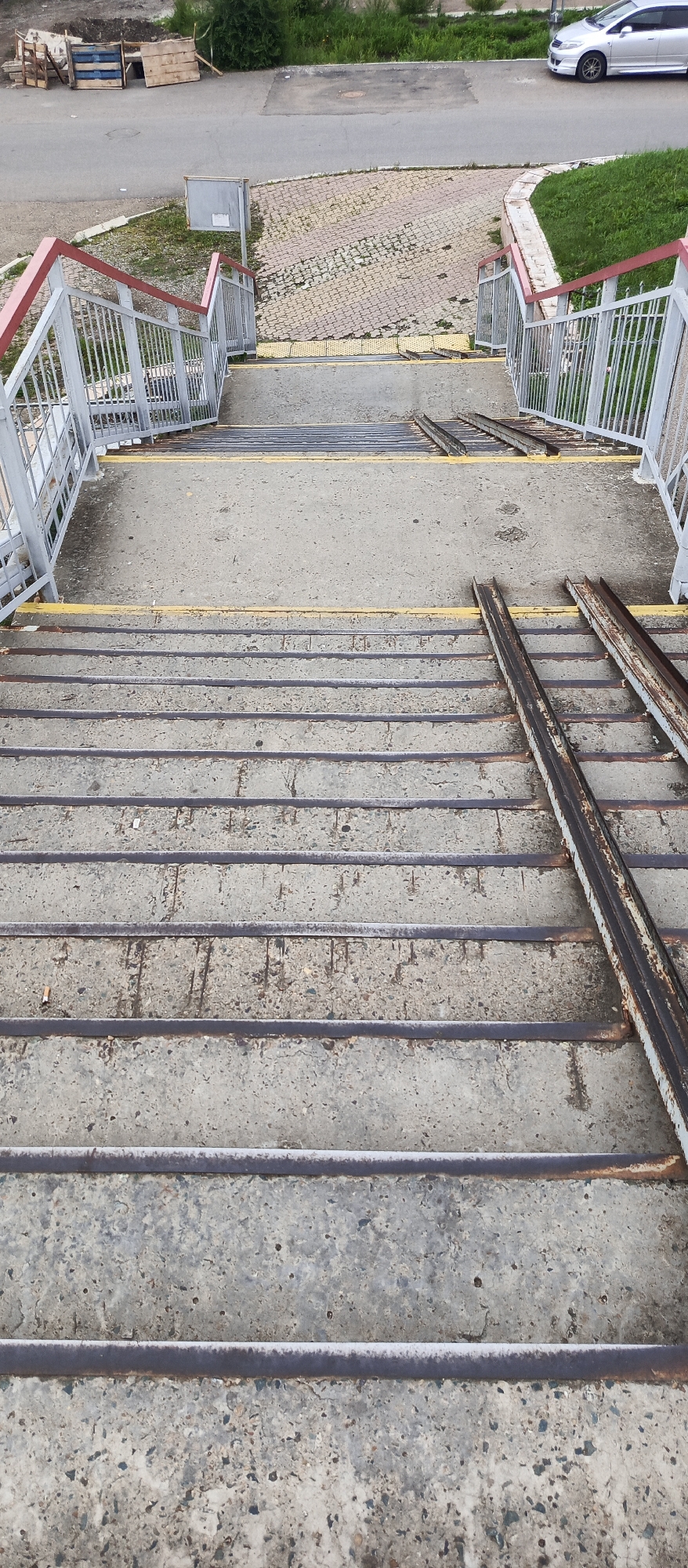 The image shows a downward view of a concrete pedestrian overpass in Pogranichny, Russia. The overpass features metal railings with red tops and yellow warning strips on the stairs. Two sets of rusty metal ramps are installed alongside the concrete stairs. The environment includes a paved road and some greenery visible at the top of the frame, with a silver car parked on the side of the road. The concrete stairs are worn and show signs of weathering and some debris.