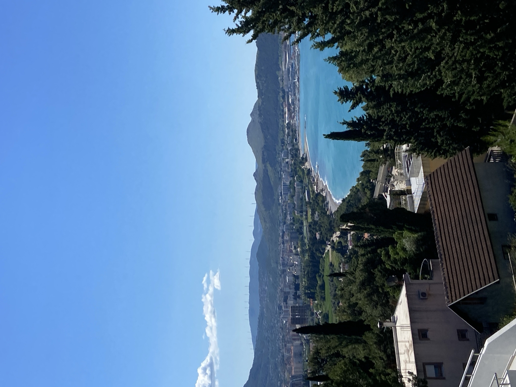 The image depicts a view of Šušanj, Montenegro. The coastline, with its beach and turquoise water, is visible along with the city stretching along the base of the mountains. Buildings of varying sizes dot the landscape. Dense green trees occupy the foreground and frame the view. The sky is clear and blue, with a few thin clouds. The buildings in the immediate foreground appear to be residential. Tall, thin trees stand among the houses and provide a contrast in height. A ridge line of mountains is visible in the background. There are what appears to be wind turbines or transmission towers on the mountainside.