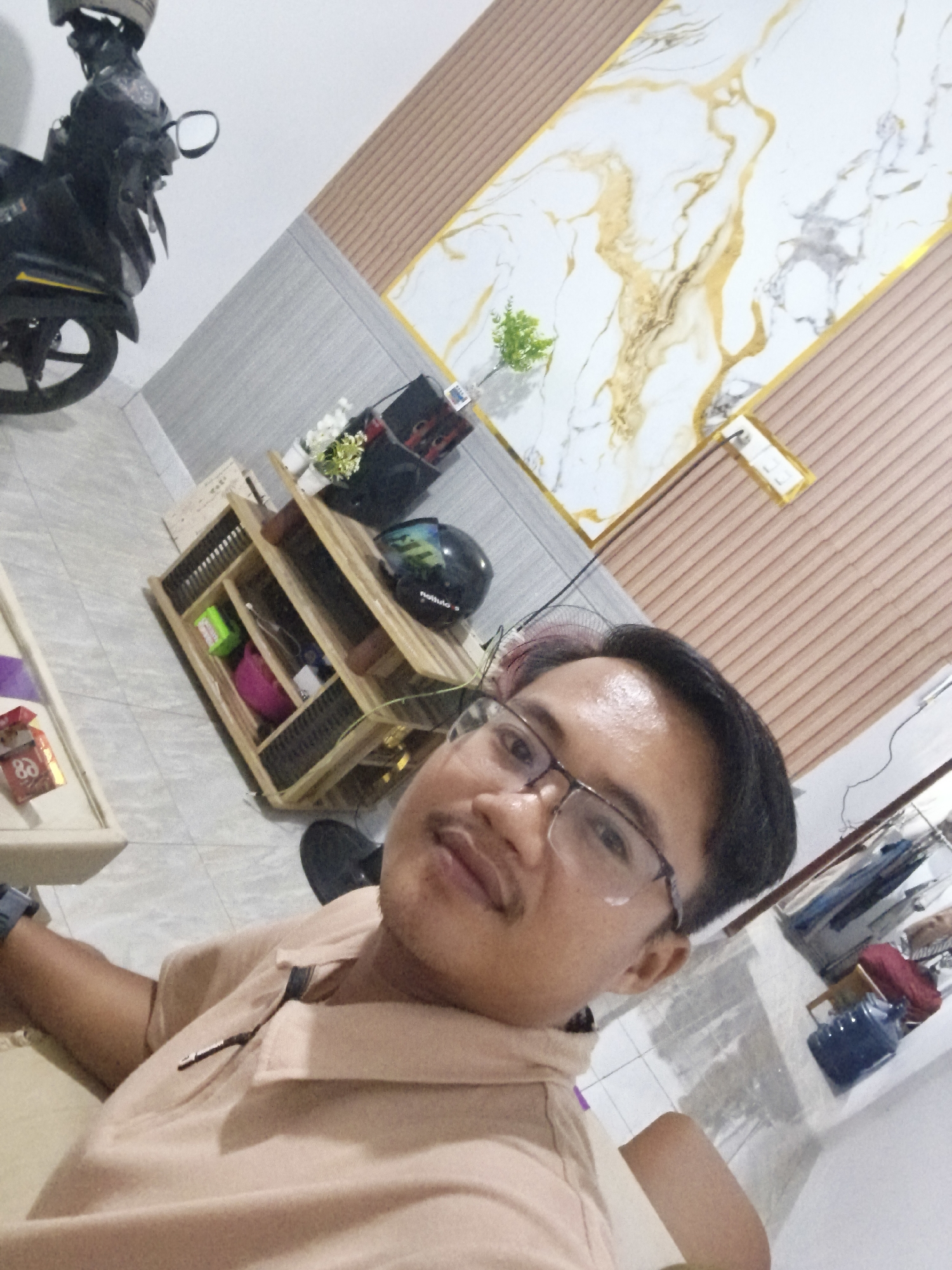 The image shows a man wearing glasses and a light brown polo shirt, looking at the camera. He is indoors, with a motorcycle visible in the background on the left. To the right of the motorcycle, there is a wooden shelf unit filled with various items, including what appears to be a helmet and some potted plants. The wall behind the shelf unit is decorated with a large, abstract art piece featuring gold and white marble patterns. The floor is tiled. The overall scene suggests a residential or personal space. This location is identified as being in Pangkalpinang, Indonesia.