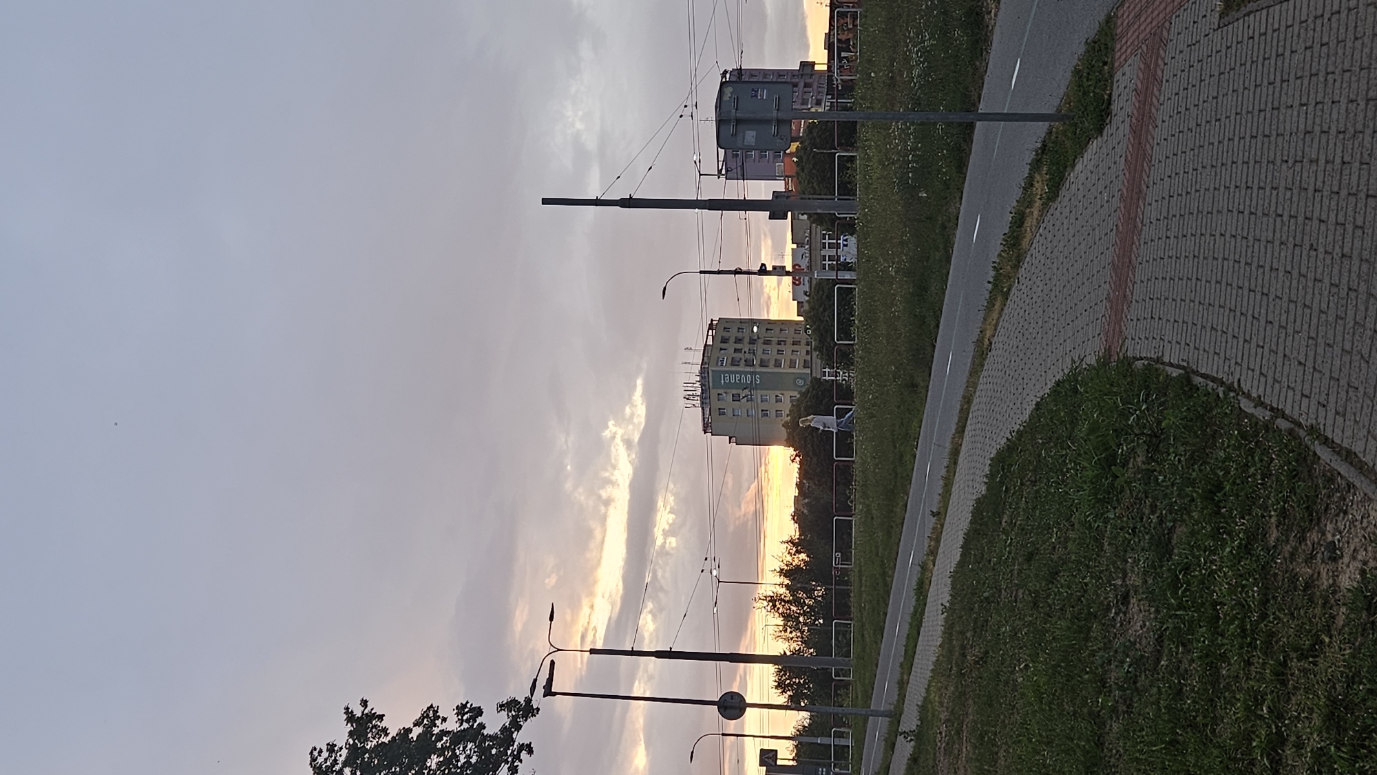 The image captures a street scene in Prešov, Slovakia, likely during a sunset due to the warm hues in the sky. A paved pedestrian walkway curves into the foreground, bordered by a grassy area and a road. In the mid-ground, a woman is walking on the sidewalk adjacent to the road. Behind her, there are several multi-story buildings, one of which prominently displays the "Slovak Telekom" logo. Utility poles and overhead wires for a tram system are visible, indicating an urban environment. The sky is a mix of grey clouds and areas of soft orange and yellow light, suggesting the sun is setting or has recently set. Trees and vegetation line the street, adding to the urban park-like atmosphere.