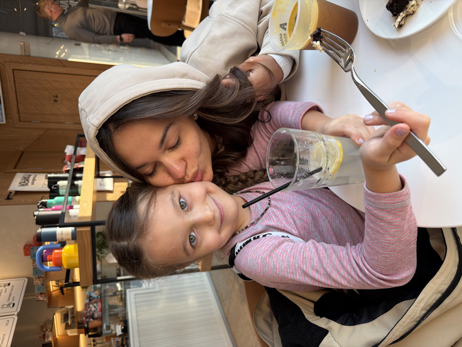 A woman in a beige hoodie kisses a smiling young girl with blue eyes, both wearing pink long-sleeved shirts. They are seated at a white table with a glass of lemonade, a drink in a plastic cup, and a piece of cake. A fork with a bite of cake is held in the girl's hand. A shelf with colorful items is visible in the background, along with a blurred figure of a man.