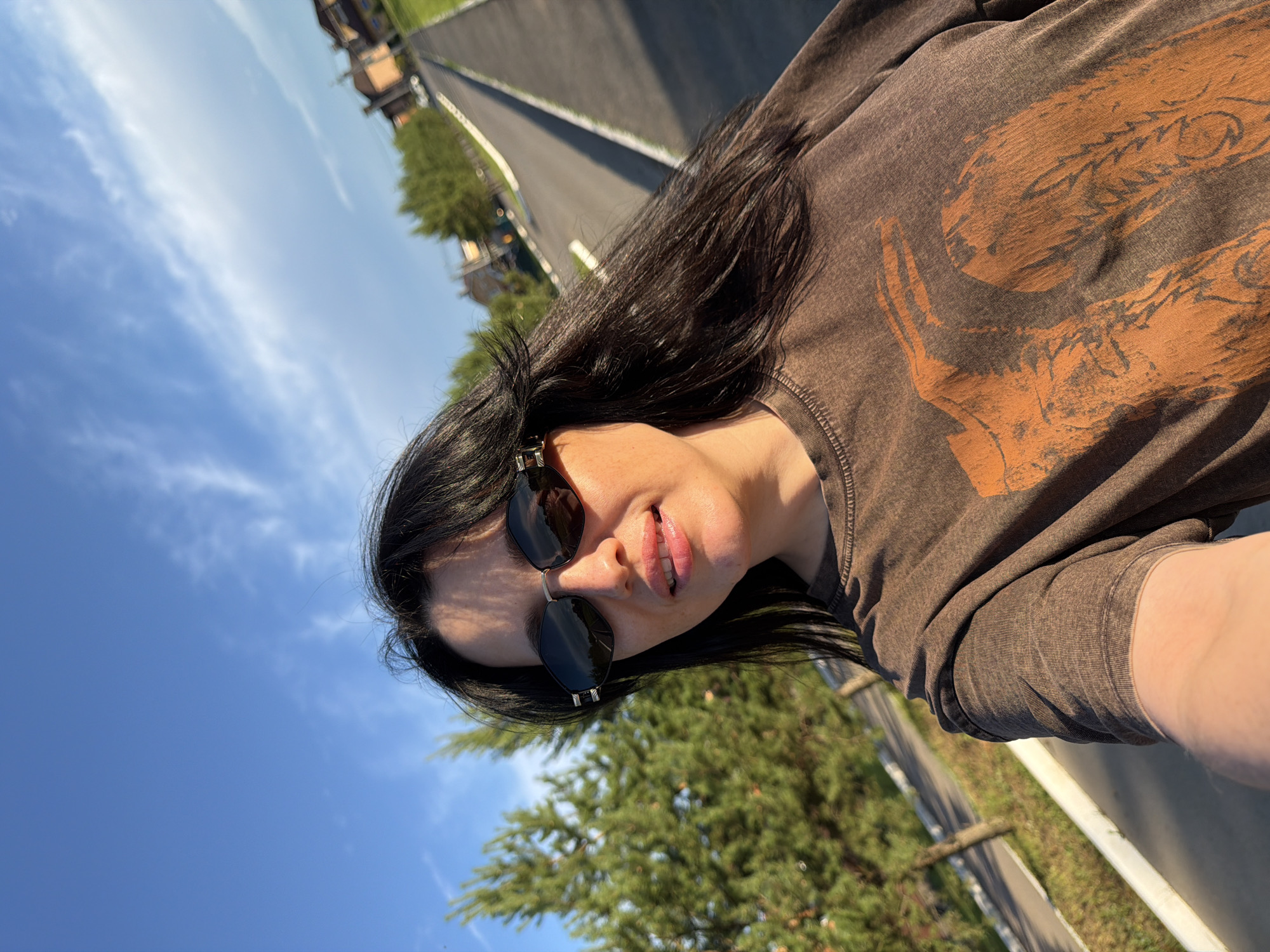 A woman is taking a selfie outdoors on a sunny day. She has long, dark hair and is wearing dark sunglasses and a brown t-shirt with an orange graphic print. She is standing on a paved area next to a grassy verge with trees lining a road that recedes into the distance. Residential buildings are visible on the right side of the road. The sky is blue with some wispy white clouds. The lighting suggests it is late afternoon. The location is the city of Shmidtovo, Russia.
