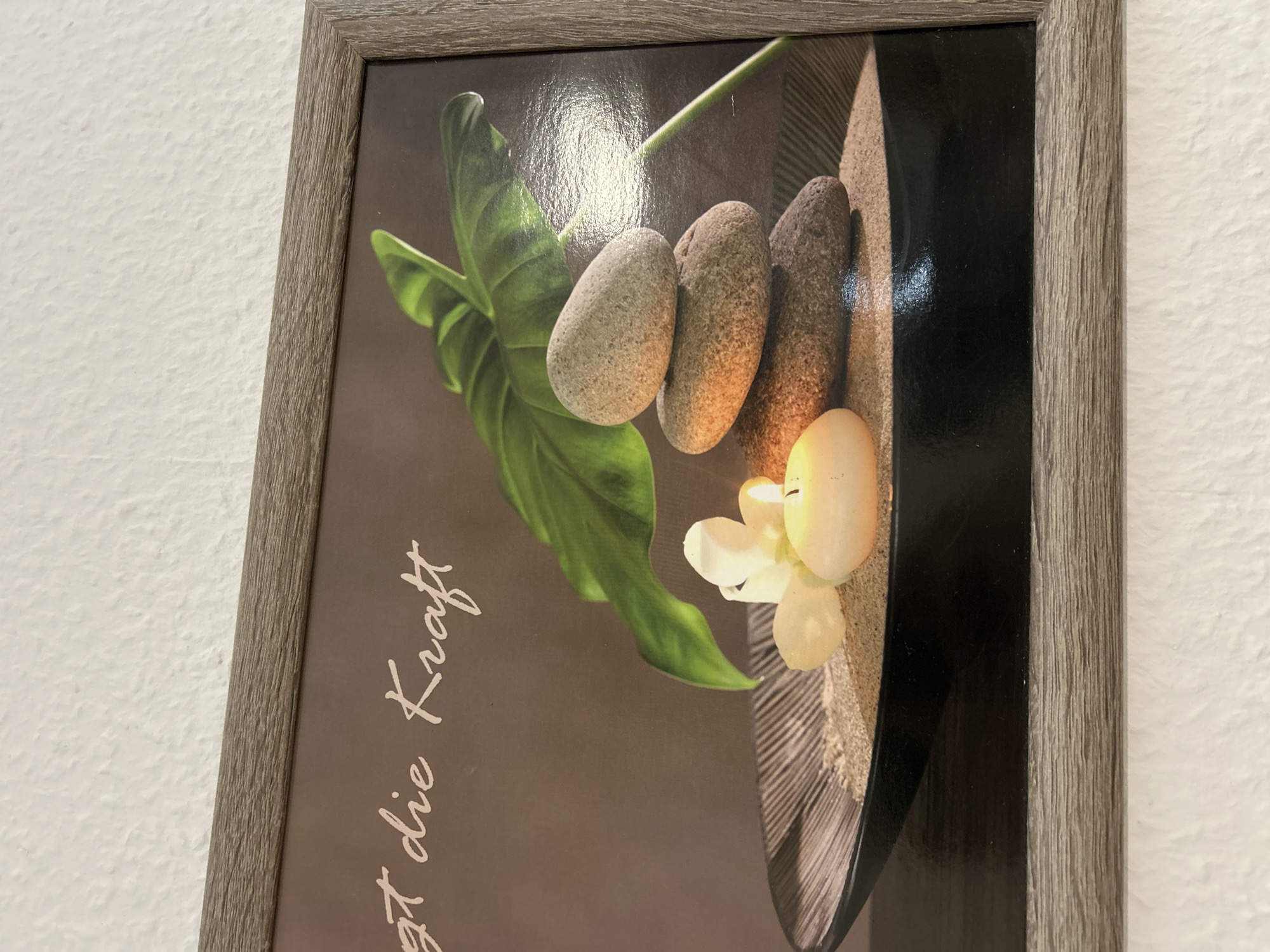 A framed picture is hanging on a textured white wall in Waldbröl, Germany. The picture is enclosed by a rectangular, light brown wooden frame with a prominent horizontal grain. Within the frame, the image displays a still life composition. In the foreground of the image, a dark, shallow bowl with subtle radial striations on its inner rim is filled with light brown sand. Positioned on the sand, to the right, is a vertical stack of three smooth, oval-shaped, greyish-brown stones, diminishing in size from bottom to top. To the left of the stone stack, a small, pale yellow, irregularly shaped candle burns with a distinct flame, casting a warm glow. Beside the candle, to its left, are two small, white, waxy flower blossoms. A large, vibrant green tropical leaf, featuring visible veins, arches downwards from the upper right quadrant of the image. The background of the picture is a plain, dark brown hue. In the upper left portion of the image, white cursive text is visible, reading "gt die Kraft". A noticeable, elongated, bright reflection is present on the glossy surface of the picture, extending diagonally from the upper right towards the center-right, indicating an external light source.
