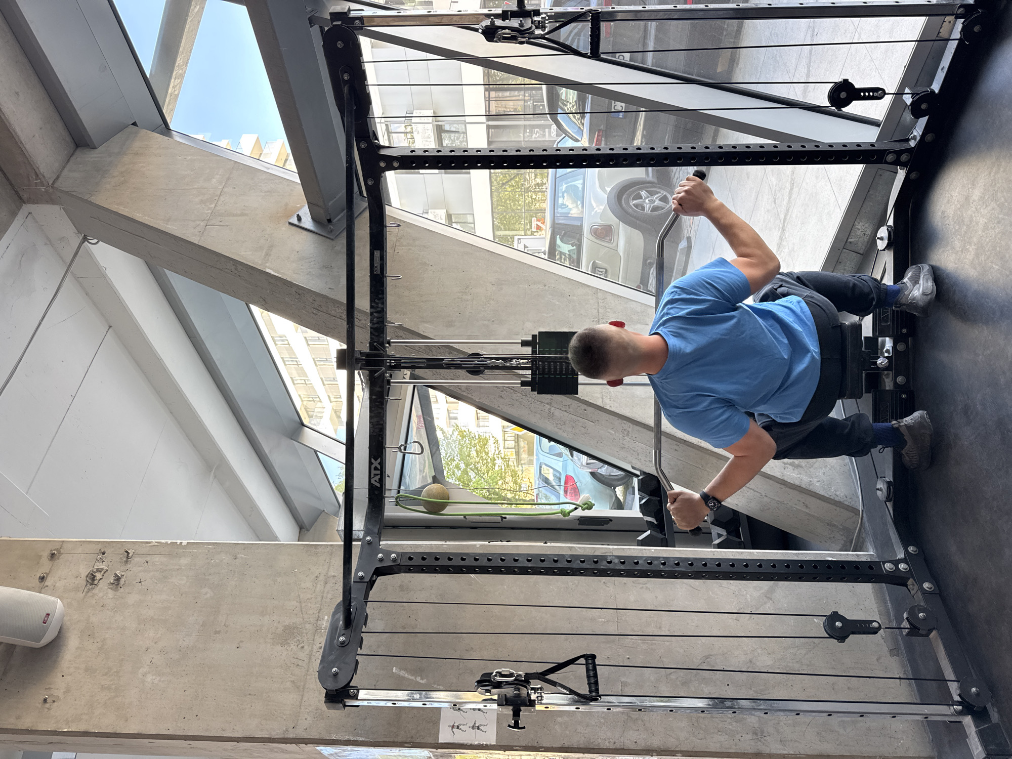 A man in a blue t-shirt and dark pants is actively engaged in a seated cable rowing exercise on gym equipment. He is positioned with his back to the camera, holding the handles of the machine and pulling the weights.
The exercise takes place in a modern gym environment, characterized by prominent concrete structural beams and large floor-to-ceiling glass windows. Through these windows, an exterior street scene is visible, including a white SUV and reflections of other buildings and greenery, indicating an urban setting. The interior design features a combination of raw concrete elements and white walls, with some grey metallic structures.
The gym equipment is a black cable machine with multiple pulleys, cables, and weight stacks. The brand "ATX" is visible on the vertical frame of the machine. At the base of the equipment, small stick figures illustrating exercise instructions are present, along with a piece of paper bearing the Cyrillic text "ТАК". A white speaker is mounted on one of the concrete walls. A yellow-green ball is secured with a green rope and a carabiner, attached to a part of the gym frame near a window.
The scene is well-lit by natural light streaming through the windows, suggesting it is daytime with a clear sky visible outside. The man is wearing a watch on his left wrist and grey athletic shoes with blue socks. The location is Sofia, Bulgaria.
