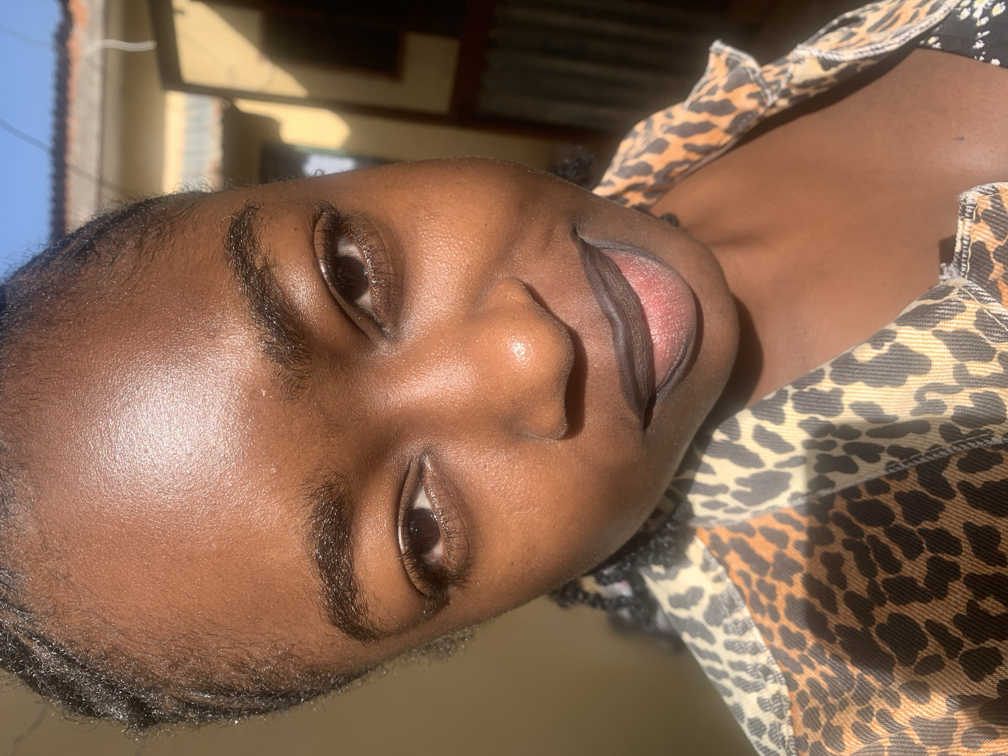 This close-up portrait, taken outdoors in Rafin Par, Nigeria, features a young person with dark skin. The subject's head is tilted, and they are looking directly at the camera with a relaxed expression, a slight smile on their face, and lips gently parted. Bright, direct sunlight illuminates their face, creating a natural sheen on their forehead, cheeks, and nose.
Visible features include dark, expressive eyes with prominent eyelashes, well-defined eyebrows, and full lips. The person is wearing a garment with a distinctive leopard print pattern, which is visible on the right side of the frame and covers part of their shoulder. The background is softly blurred, showing hints of a clear blue sky and simple building structures, indicating a daytime setting. No text is visible in the image.