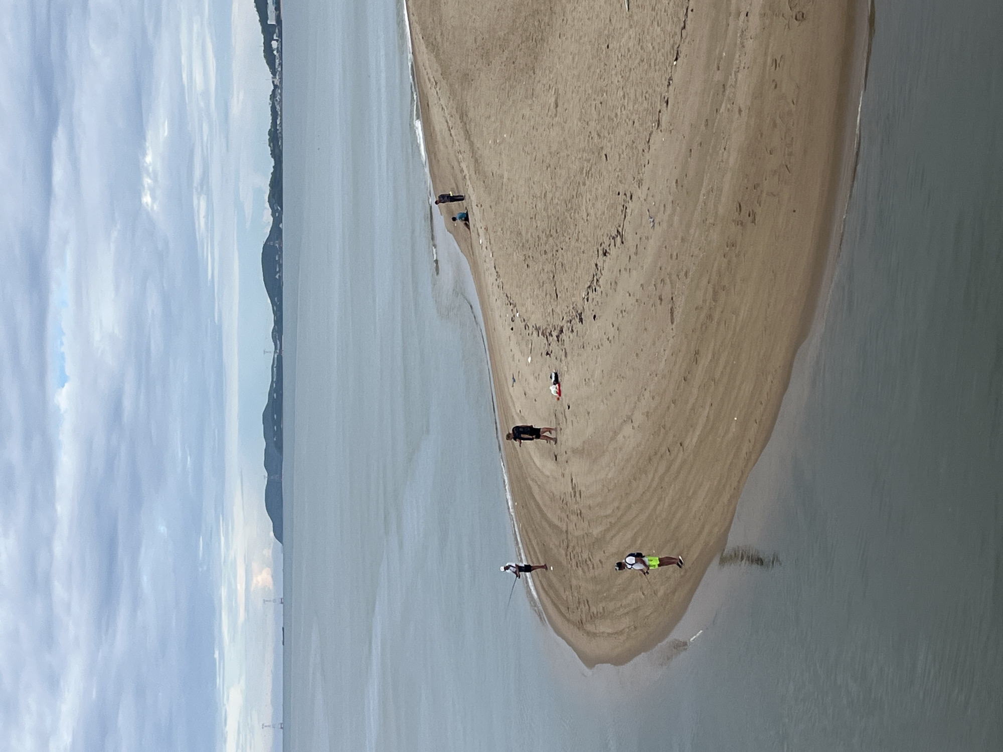 Captured aerially in Bắc Nha Trang Ward, Vietnam, the scene presents a sandy peninsula surrounded by water. Several people are dispersed along the beach's edge, some appear to be fishing. The sky is overcast with gray clouds. Distant mountains are visible across the water. A bridge-like structure stretches across the water in the background.