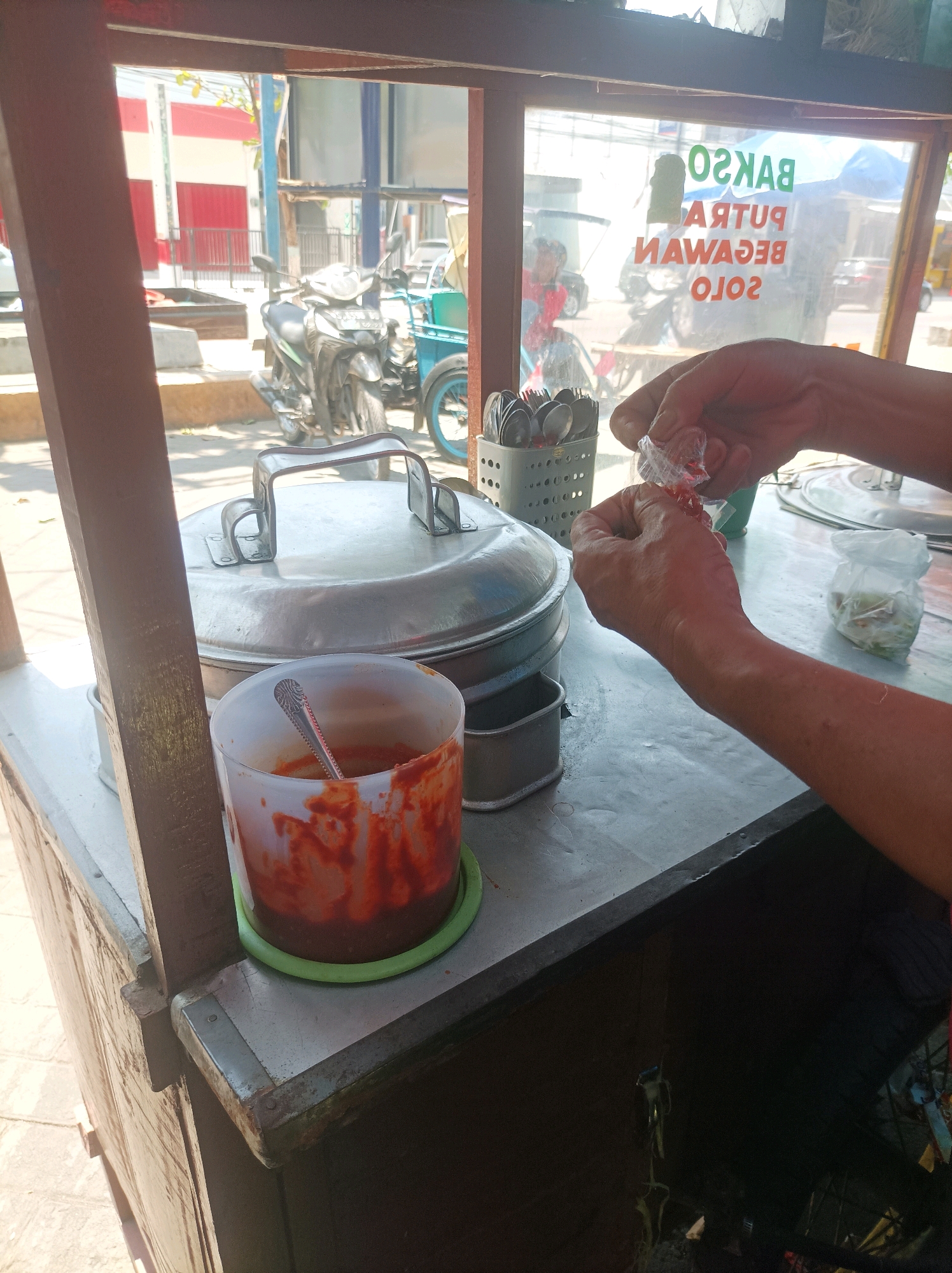 The image depicts a close-up view of a food cart, likely selling meatballs, in Sumuragung, Indonesia. The cart features a large, silver, lidded pot in the foreground, with a clear plastic container filled with a red sauce and a spoon to its right. To the left of the pot, a metal basket holds an assortment of spoons. In the background, visible through the cart's glass, is a street scene with motorcycles and buildings, one with red and white striped walls. A person's hands are prominently featured in the center-right of the frame, actively opening a small, clear plastic package containing red items, possibly condiments or chili sauce. The overall environment appears to be outdoors on a sunny day, with harsh shadows indicating bright sunlight. The lettering on the glass of the cart, reversed from the viewer's perspective, indicates "BAKSO PUTU BEGAWAN SOLO."