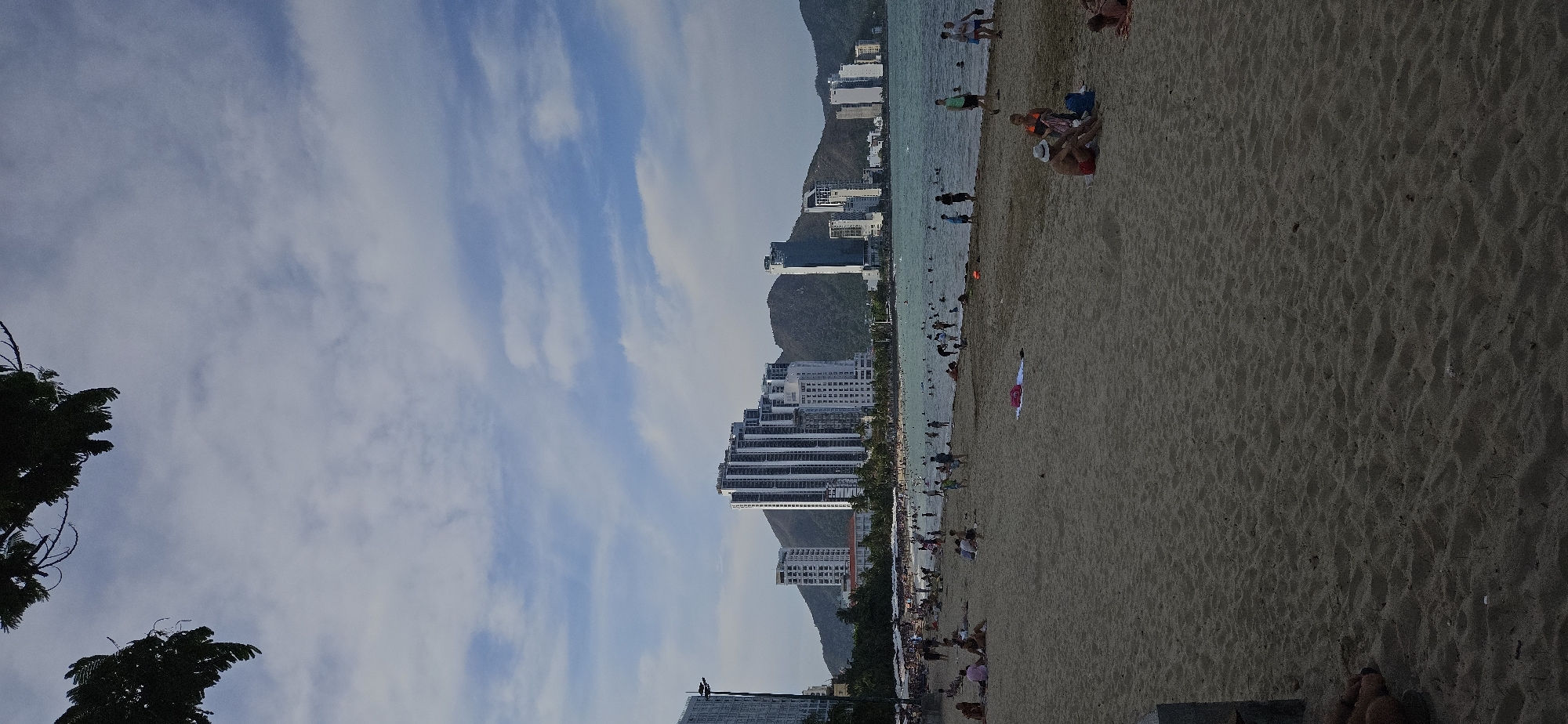 The image depicts a beach scene in Bắc Nha Trang, Vietnam. The sandy beach is crowded with people swimming, sunbathing, and walking along the shore. The water is clear and shallow. In the background, tall buildings line the coast, framed by mountains. The sky is blue with scattered clouds. Trees are located on the left side of the image.
