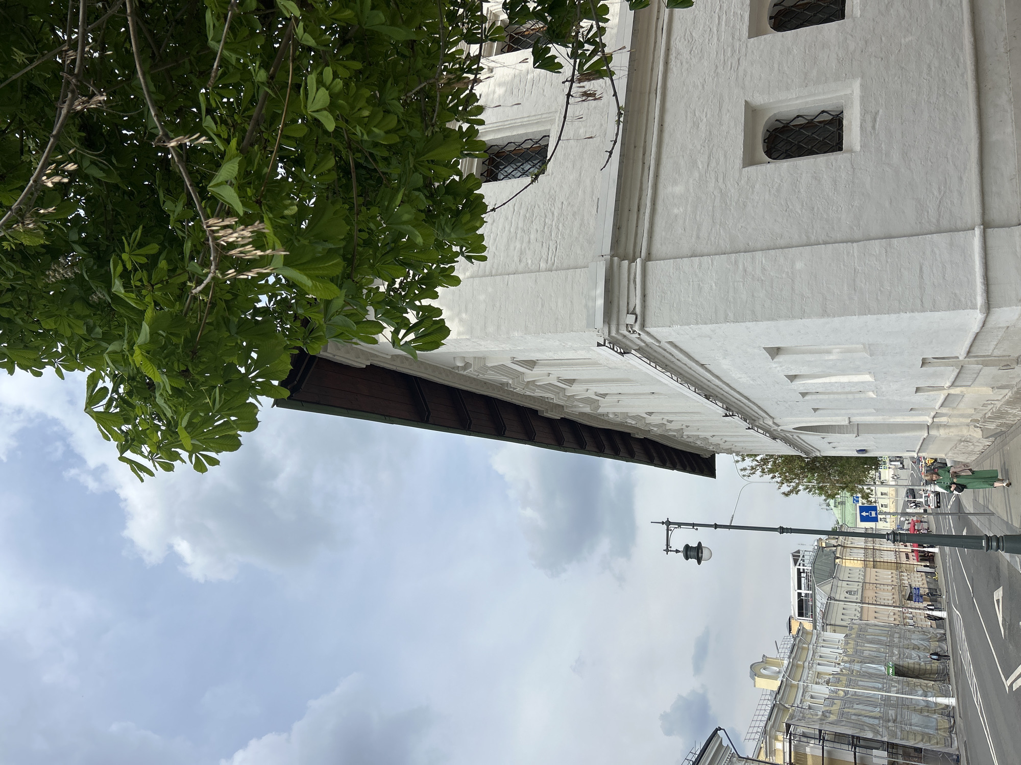 A street view in an unidentified city shows a white building on the left, partially obscured by green foliage. The building features a brown roof overhang and barred windows. The sky above is partly cloudy. A street with lampposts and various buildings runs horizontally across the lower part of the image. Pedestrians can be seen walking on the sidewalk.
