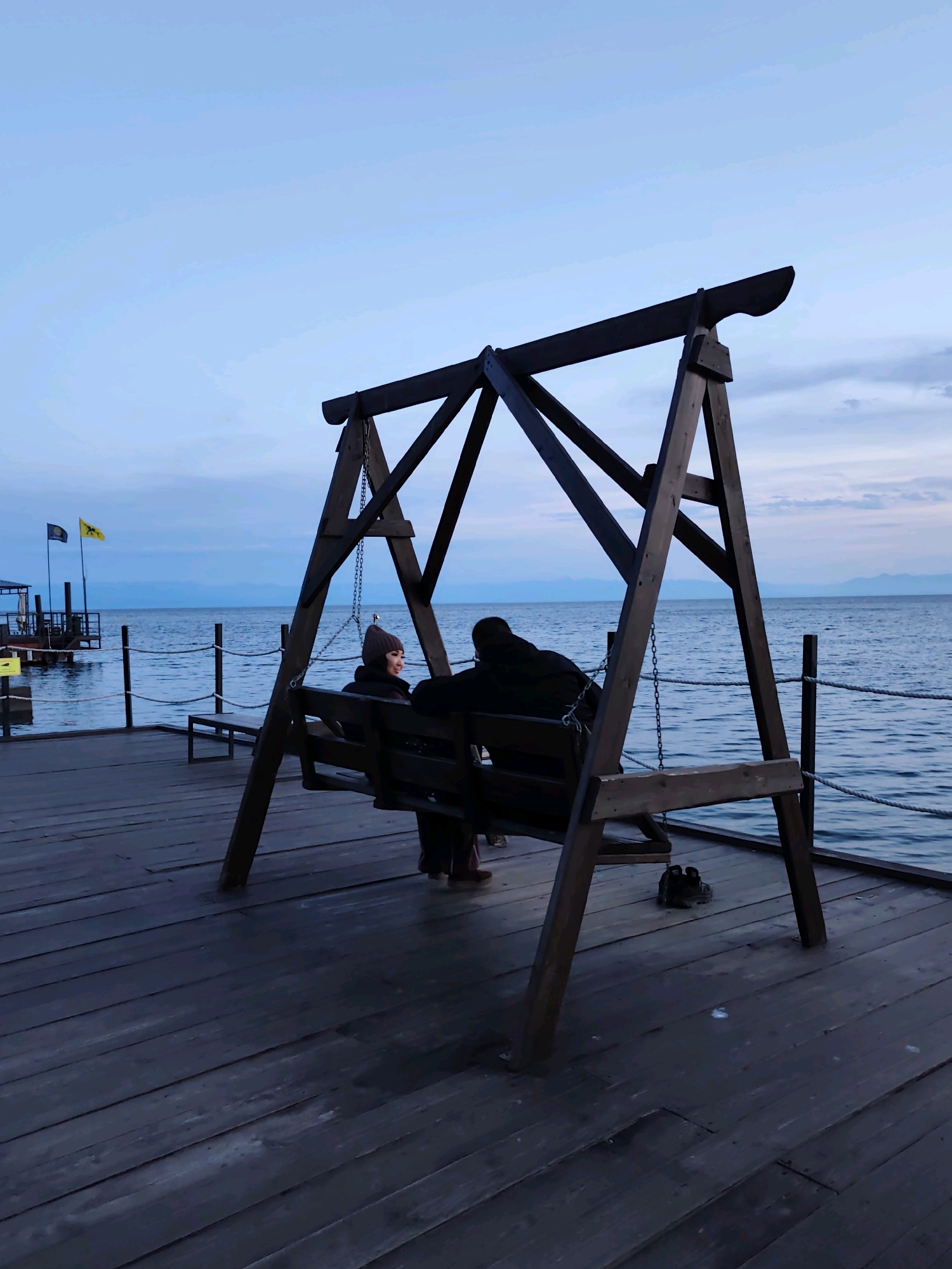 Two individuals, one facing towards the viewer and the other away, are seated on a wooden swing located on a wooden pier. The person facing the viewer is wearing a beanie and a jacket, and appears to be smiling. The other individual is wearing a dark jacket. The swing is suspended by chains from a wooden A-frame structure. The pier extends towards a body of water, likely Lake Baikal, with mountains visible in the distance. Two flags are planted on a structure further down the pier. The sky is overcast, and the overall lighting suggests it is dusk. The scene is set in Listvyanka, Russia.