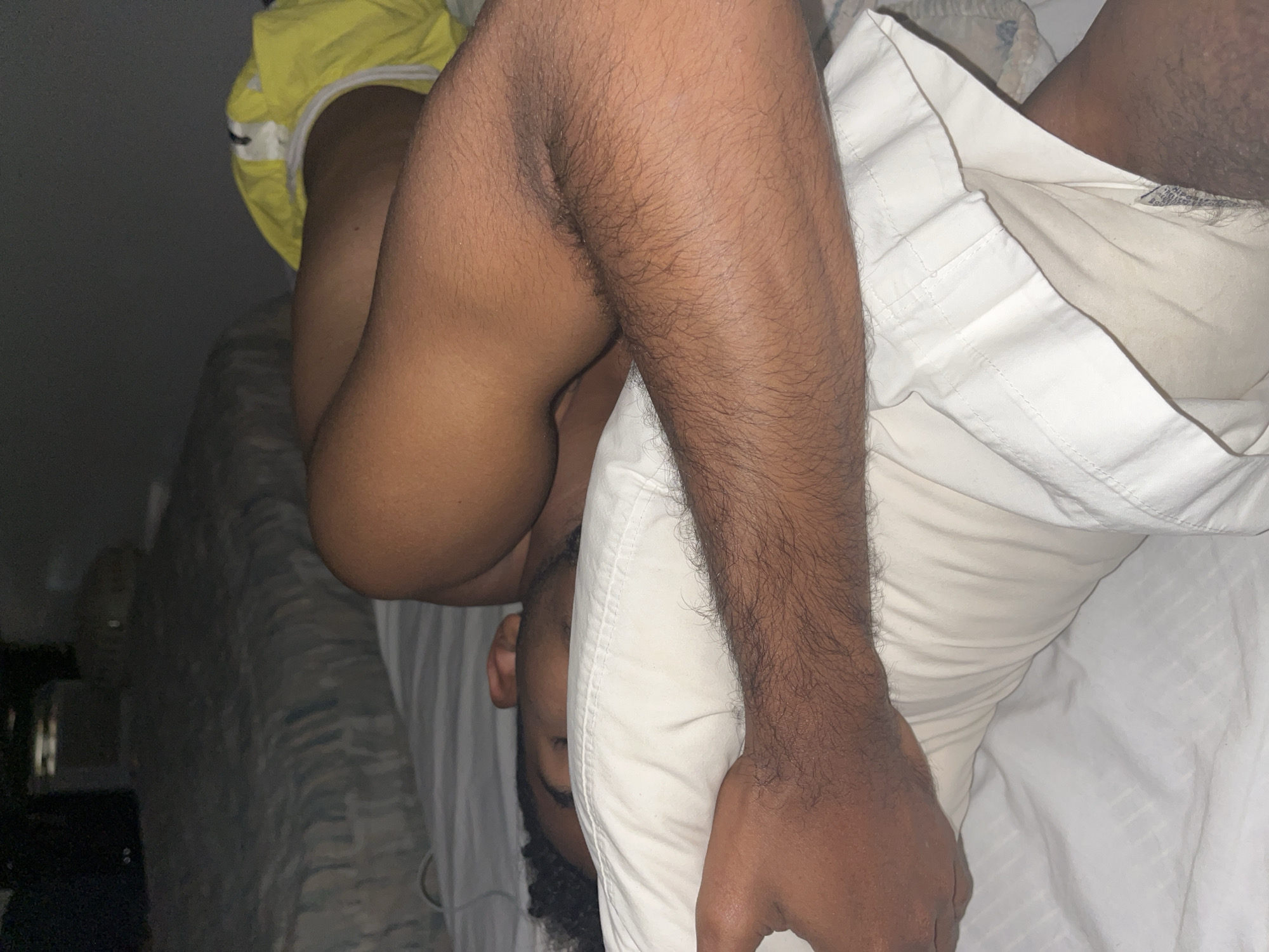 A person, appearing male due to body hair and visible stubble, is depicted lying on their left side on a bed, seemingly asleep or resting. Their head, adorned with dark, curly hair, rests on a white pillow, partially obscuring their face, though the top of their head and a portion of their right ear are visible. One arm, covered in dark hair, is bent and positioned across the pillow near their head. The person is wearing light-colored, possibly white, shorts. A bright yellow garment is bunched near their upper back/shoulder area on the bed. The bed is covered with white sheets and a white pillow with subtle horizontal stripes. In the dimly lit background, another bed with a patterned bedspread (grey, blue, and white tones) is visible, along with indistinct dark objects. The image does not contain any discernible elements that specify its location within Porlamar, Venezuela, or any other geographical area.