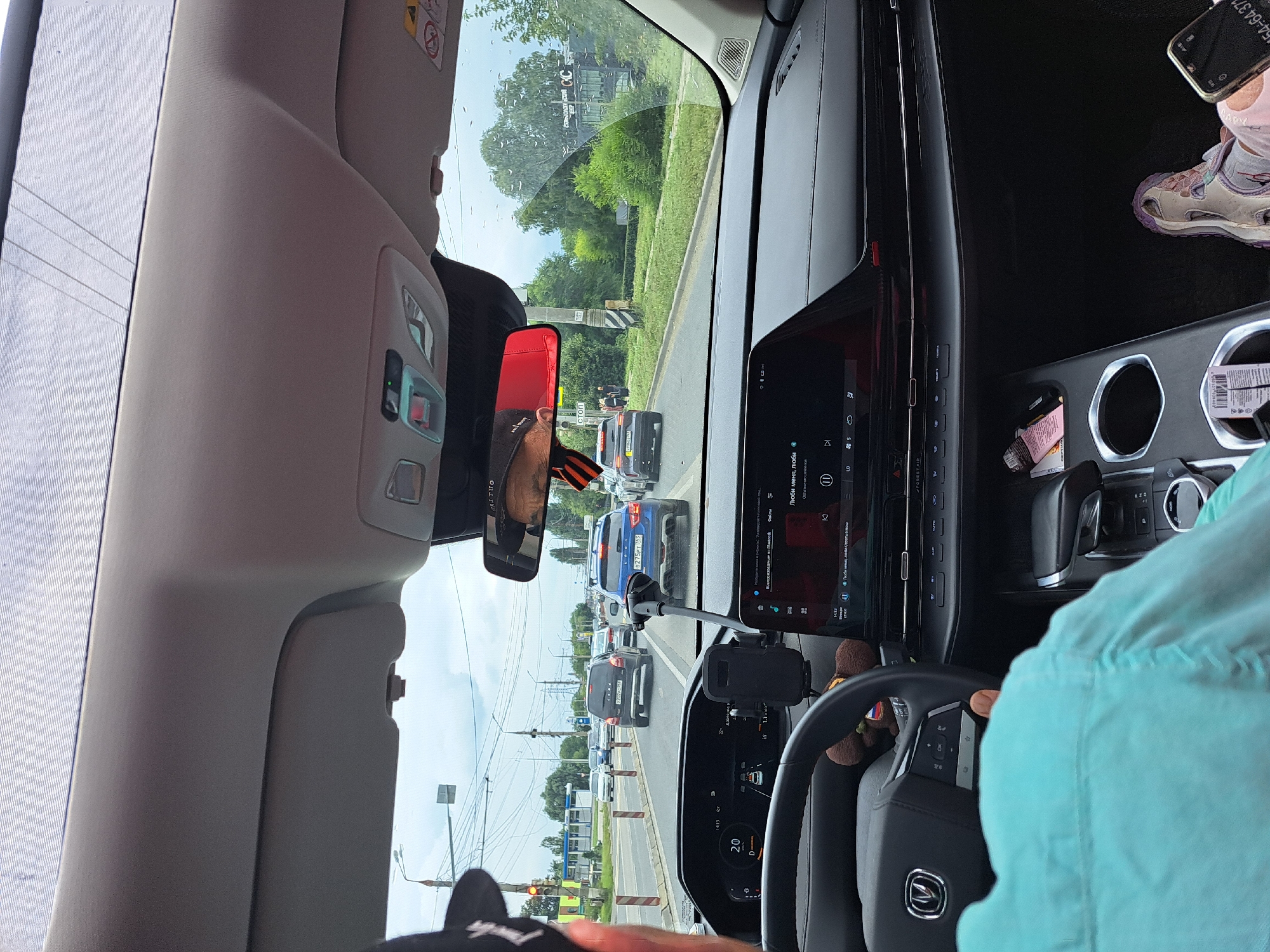 The image shows the interior of a car as viewed from the passenger seat, with the driver's seat partially visible. The car is driving on a road in what appears to be a city, with other vehicles on the road and buildings visible in the background. The sky is overcast.
In the foreground, the dashboard of the car is prominent, featuring a large infotainment screen displaying what appears to be music controls and a navigation interface. The steering wheel is also visible, with a stuffed toy hanging from it. A person's arm wearing a light blue shirt is visible on the left side of the frame, suggesting they are either the passenger or the driver.
The car's windshield offers a view of the road ahead, where several cars are driving. Traffic lights and street signs are also visible. On the right side of the dashboard, a person's legs are visible, wearing light-colored shoes. A smartphone is also visible in the lower right corner.
The overall impression is a snapshot of a car journey in an urban environment, likely during the daytime. The specific location is not identifiable from the provided image.