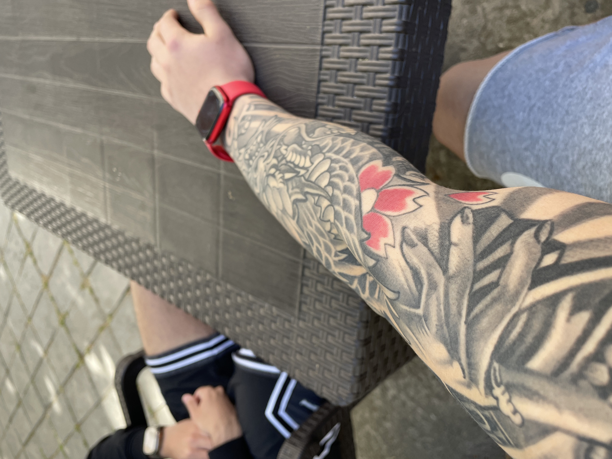 An overhead shot captures an outdoor scene in Plovdiv, Bulgaria, featuring two individuals seated at a dark grey, textured outdoor table, likely made of rattan.
The main subject is a person's arm, heavily adorned with an intricate, full-sleeve Japanese-style tattoo. The tattoo predominantly features grey-toned scales and patterns, possibly depicting a dragon or mythical creature, interspersed with vibrant red cherry blossom flowers. A prominent red Apple Watch is worn on the wrist of this arm, which rests on the table.
In the background, the lower half of another person is partially visible, wearing light grey shorts. In the lower left corner of the image, the legs of a second person are seen, clad in black track pants with distinctive white stripes, and a light-colored watch is visible on their wrist. The ground beneath the table is paved with light-colored, patterned stones, indicative of an outdoor patio or garden area.
The scene is brightly illuminated by natural light, suggesting it is daytime. The people appear to be relaxing or socializing at the table. No discernible text is visible on any of the watches, clothing, or the table surface.