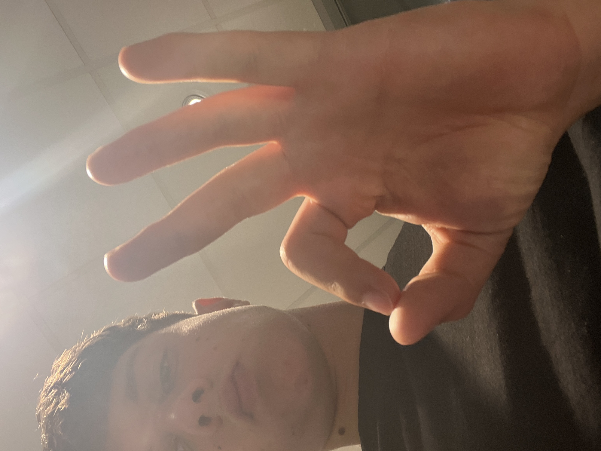 A selfie taken from a low angle. A young man with short brown hair and visible acne is looking down at the camera. His mouth is slightly open. He is wearing a black shirt. His right hand is raised, making an "OK" sign with his thumb and forefinger, and extending the other three fingers. The background is a white ceiling with a recessed light fixture. The location is not evident in the image.