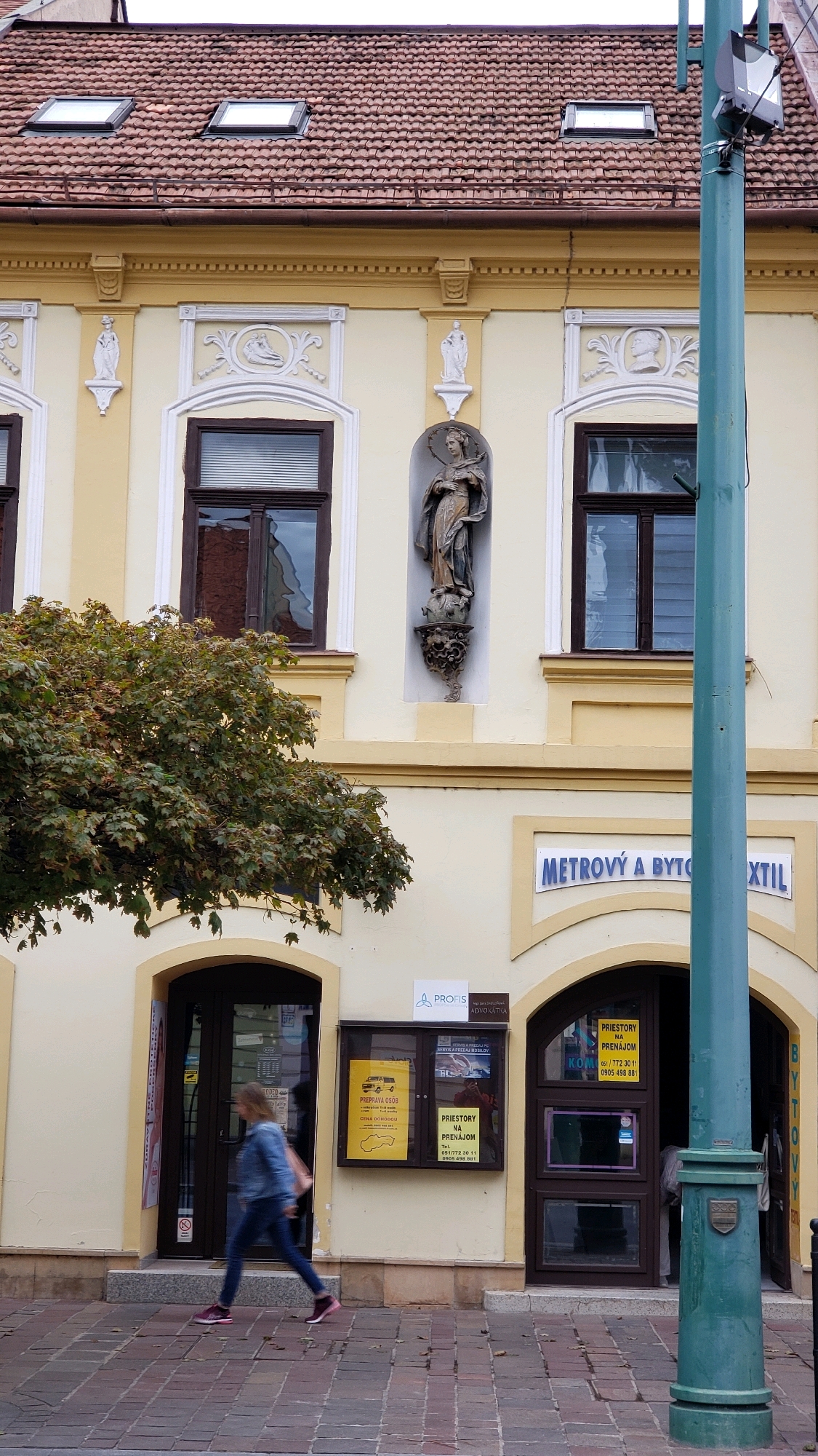 A medium shot captures the facade of a building in Prešov, Slovakia, on a bright, overcast day. The building's upper floor features a reddish-brown tiled roof with two prominent dormer windows. The yellow facade is adorned with decorative white plasterwork above two dark-framed windows on this level, and a statue of a robed figure is set within a niche between them. Below, the ground floor has a wide arched entrance with a dark-framed door and a large window. Adjacent to this is another arched entrance with a similar door and window. A green lamppost stands prominently in the foreground, to the right of the building. In front of the building, a woman in a denim jacket and blue jeans walks from left to right, her face obscured. A tree with green and brown leaves partially obscures the left side of the building. Signage on the building includes "METROVÝ A BYTO" and "TEXTIL" in blue lettering above an arch, and a professional office sign that reads "A PROFIS ADVOCATKA". A bulletin board displays various notices, including one with a yellow background and an image of Slovakia. The ground is paved with reddish-brown cobblestones.