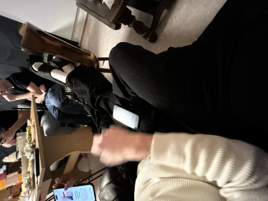 A group of people gather around a table. A person in a white sweater is holding a phone with an article displayed. Another phone rests on black pants. An individual wears black slides with white socks, feet propped on a footstool. A dark-haired person sits with arms crossed. A table with food and drinks is present. A dark wooden side table is visible in the background.
