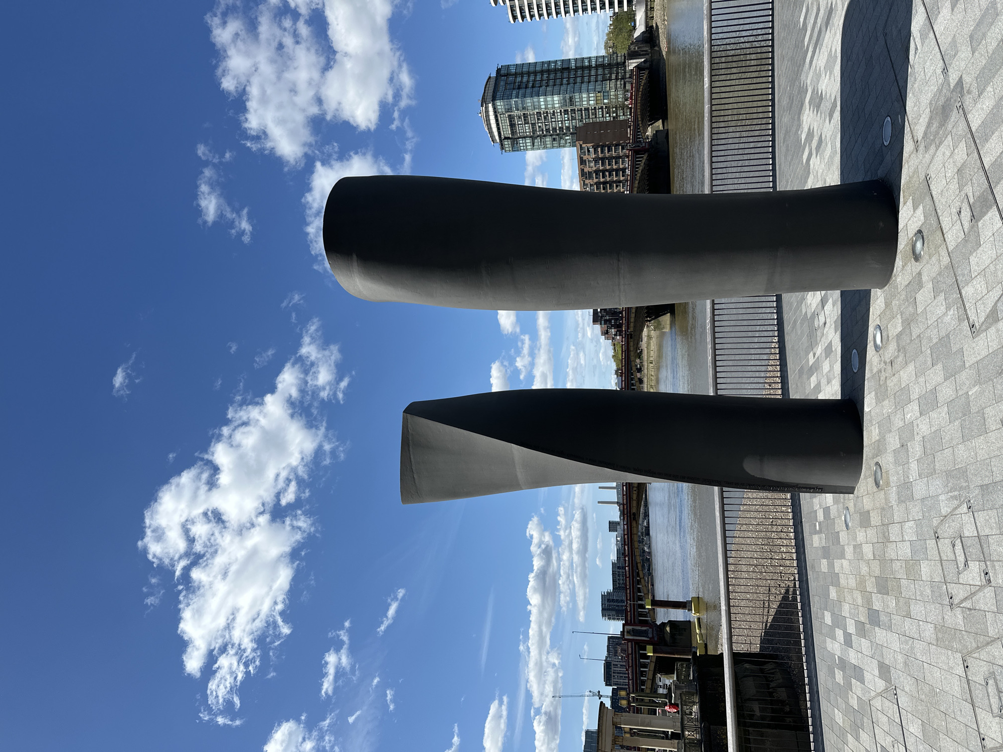The image depicts two dark gray sculptures, likely made of metal, positioned on a gray brick-paved walkway next to a river in London, United Kingdom. In the background is a building with glass windows, several other buildings, a bridge crossing the river, a metal fence, and other architecture characteristic of a cityscape. The sky is blue with scattered white clouds. There are no people visible in the image. The sculptures appear to be modern art installations.