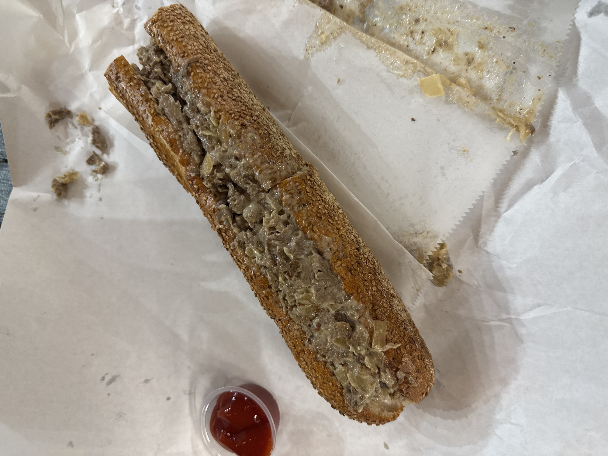 A close-up, high-angle shot depicts a cheesesteak sandwich on a white paper-lined surface, potentially from a restaurant or takeout setting. The sandwich features a sesame seed-covered roll filled with chopped meat, possibly cheese, and onions. Adjacent to the sandwich is a small, clear plastic container holding a red sauce, likely ketchup. There's also a partially visible piece of white paper with some greasy residue, suggesting it was part of the sandwich wrapping. Loose pieces of meat are scattered around the sandwich. Based on the available information, a person or entity with knowledge of regional dishes and local establishments would need to provide specific details regarding the location.