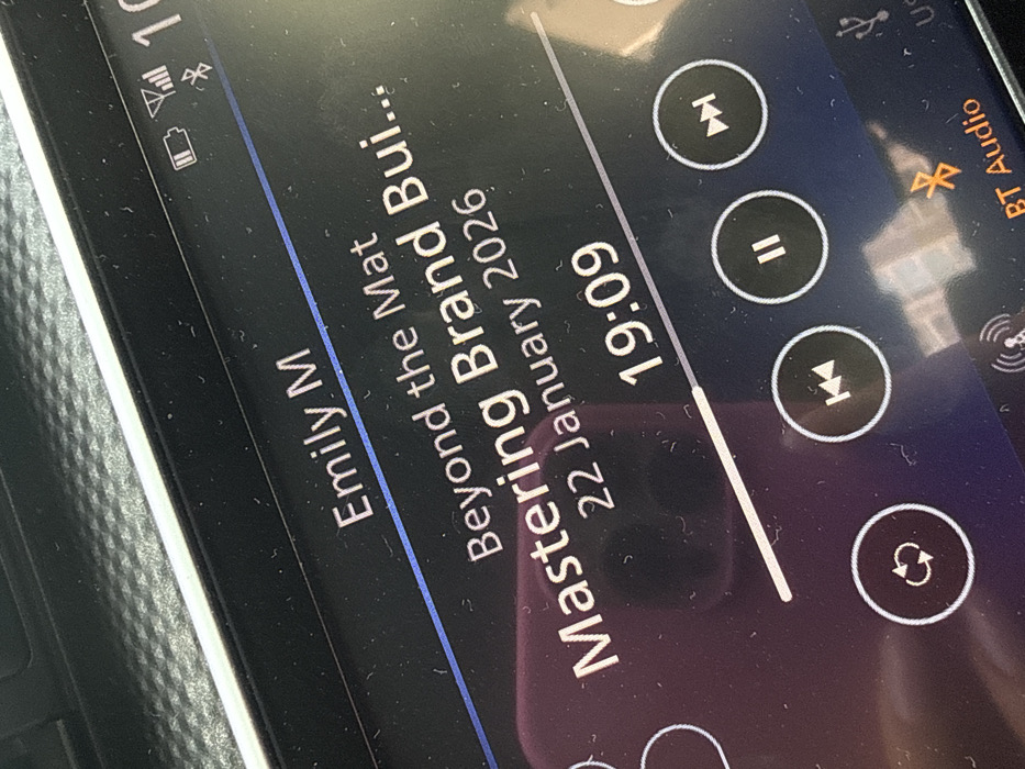 A car infotainment system shows audio playing from "Emily M" titled "Beyond the Mat" and "Mastering Brand Building" dated January 22, 2026. The playback is at 19:09. Playback controls are visible: previous track, pause/play, next track, and repeat. BT Audio is connected. A battery icon and signal strength are displayed in the top left. A USB connection is on the top right.