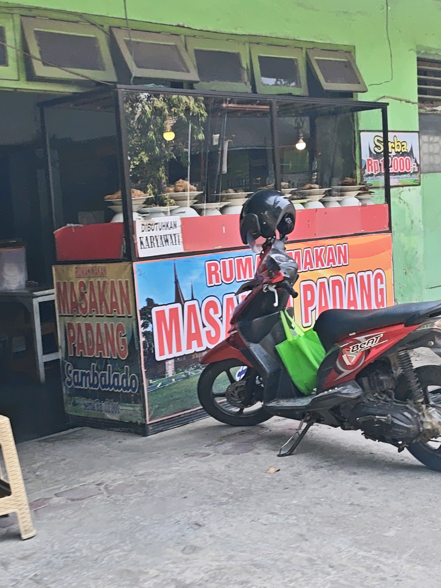 An exterior medium shot captures a street-side food stall, identified by signage as "Masakan Padang," located in Kediri City, Indonesia. The stall features a glass display case filled with various food dishes, and a red counter below. Behind the counter, a white sign reads "DIBUTUHKAN KARYAWATI," indicating a need for female employees. A red motorcycle is parked in front of the stall, with a black helmet resting on its handlebars and a green reusable shopping bag hanging from the front. The background shows a green building with windows and a sign advertising "Serba Rp1.000." The scene appears to be an ordinary day in a bustling urban environment, with the focus on the food stall and the parked motorcycle.