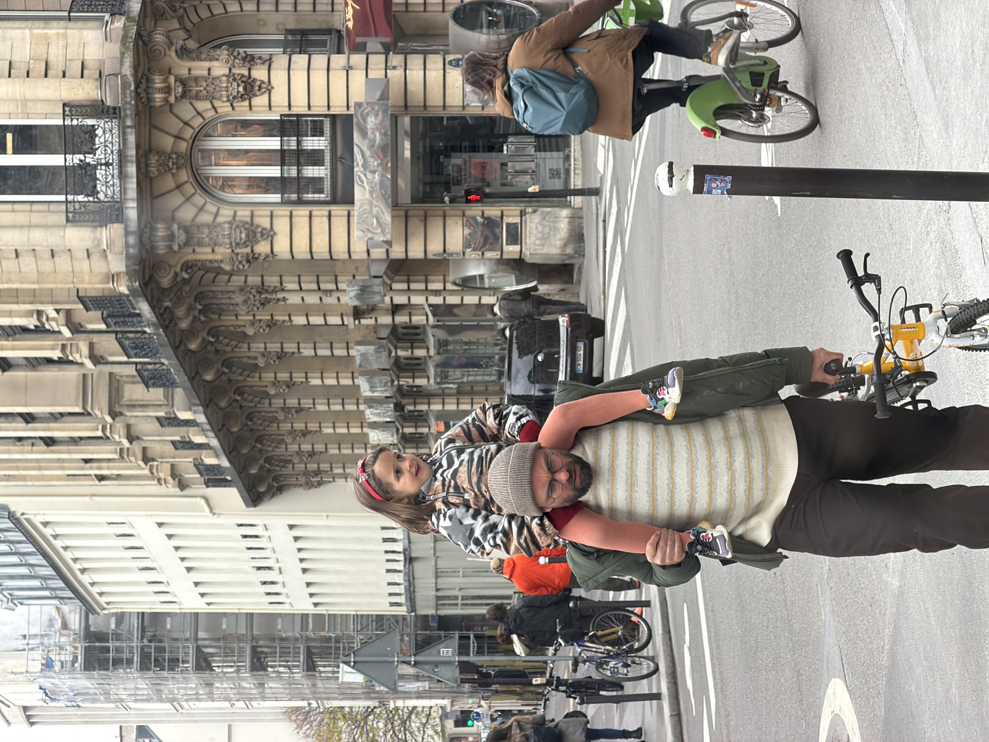 A vertical medium shot shows a man giving a young child a piggyback ride in Paris, France. The man wears a gray beanie, glasses, a light-yellow-and-white-striped sweater, a dark green jacket, and dark-colored pants. The child, who has long brown hair and a red headband, wears a gray-and-white-zebra-striped jacket with a large black star and pink long-sleeved shirt. Both wear blue sneakers with white soles. The man is on a yellow bicycle, facing to the right of the frame. The building on the left side of the frame has white siding with horizontal lines. The building on the right side of the frame has ornately carved stonework above the ground floor. Another person is riding a green-and-gray bicycle in the background, with a blue backpack. Other people, bicycles, and cars are visible in the background. A black metal street post is in the upper right side of the frame.