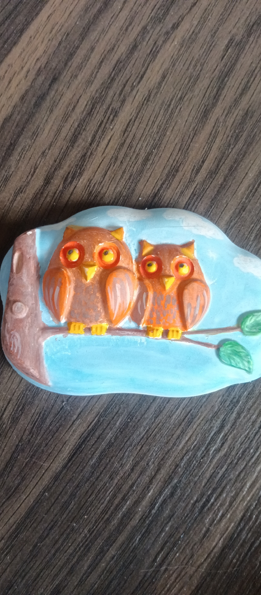 The image displays a decorative, sculpted plaque featuring two owls perched on a tree branch, set against a light blue sky with white cloud shapes. The plaque itself has an irregular, cloud-like outline.
The main subjects are two owls, depicted in a raised relief style. The owl on the left is slightly larger than the one on the right. Both owls are primarily reddish-brown and orange, with patterned feather details suggested through color variations. They both have large, round, prominent orange eyes with bright yellow centers and small, dark pupils, giving them a wide-eyed appearance. Their beaks and clawed feet are yellow.
The owls are sitting on a brown branch, which extends from a thicker, darker brown tree trunk on the far left of the plaque. A few simple green leaves sprout from the branch on the right. The background of the scene within the plaque is a uniform light blue, representing the sky, with soft white shapes suggesting clouds. A very thin, horizontal hairline crack is visible across the blue sky background, running above the head of the smaller owl.
The plaque is placed on a dark brown wooden surface, which features a distinct diagonal grain pattern. There is no discernible activity taking place, as the image captures a static, decorative scene. The artwork appears handmade or crafted, possibly from ceramic, clay, or resin, and is painted with vibrant colors. No text is visible in the image. The setting depicted is an artistic rendition of a daytime outdoor scene.