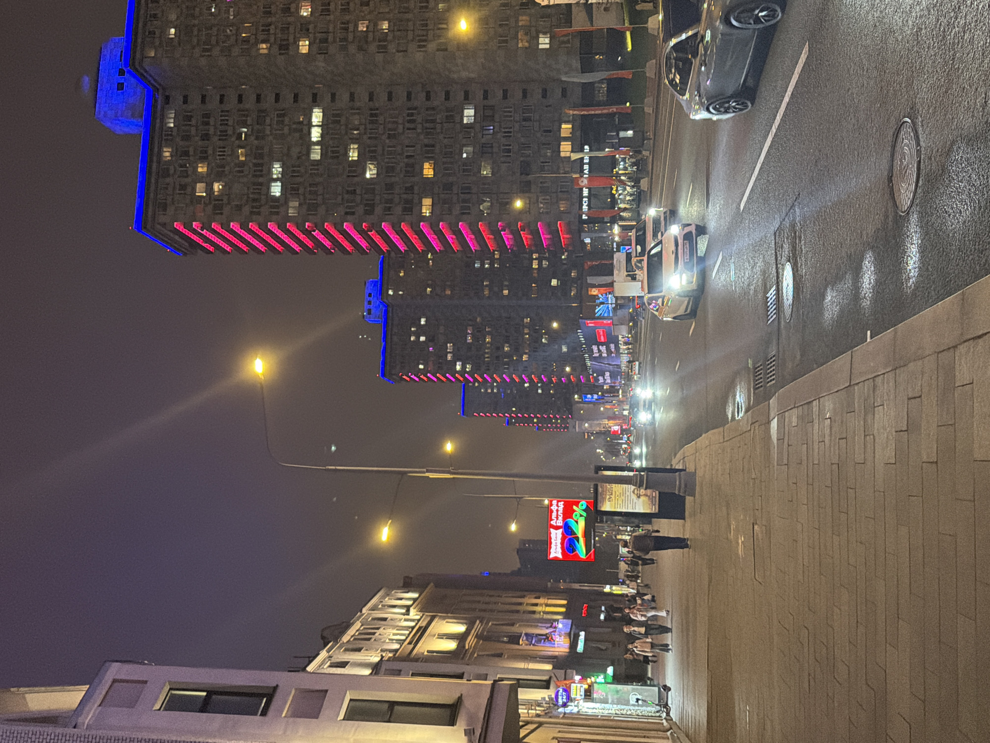The image shows a nighttime urban street scene in Moscow, Russia. Several high-rise buildings are visible, illuminated with rows of pink and blue lights. A streetlight casts a bright glow on the scene. Cars are moving along the wet asphalt road. On the sidewalk, pedestrians are walking. A large advertising billboard with colorful graphics and Cyrillic text is visible. The pavement is made of light-colored stone blocks.